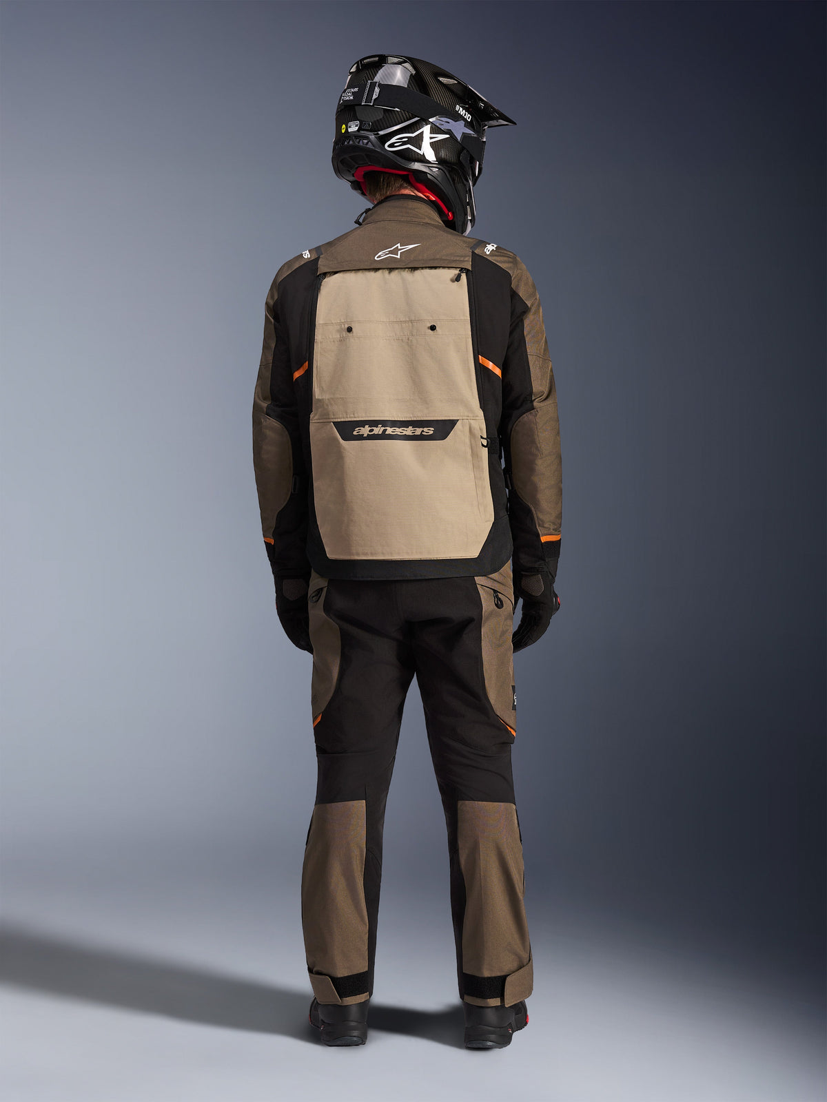 Pilota visto da dietro con giacca Alpinestars Cusco Drystar® in Walnut Aluminum Black, caratterizzata da una grande tasca posteriore color alluminio e accenti arancioni, pantaloni adventure coordinati, casco off-road nero e guanti, abbigliamento da moto in tessuto resistente per il touring.