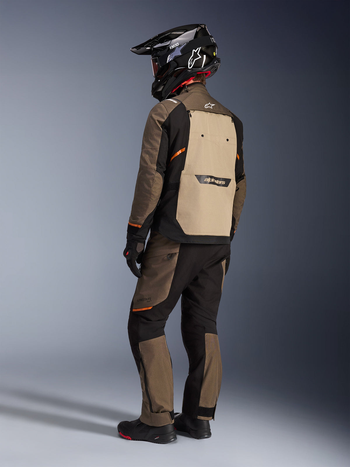 Giacca Alpinestars Cusco Drystar®, giacca adventure touring, Walnut Aluminum Black, vista posteriore che mostra il pannello posteriore marrone chiaro e i dettagli arancioni, indossata con pantaloni coordinati e casco nero in fibra di carbonio.