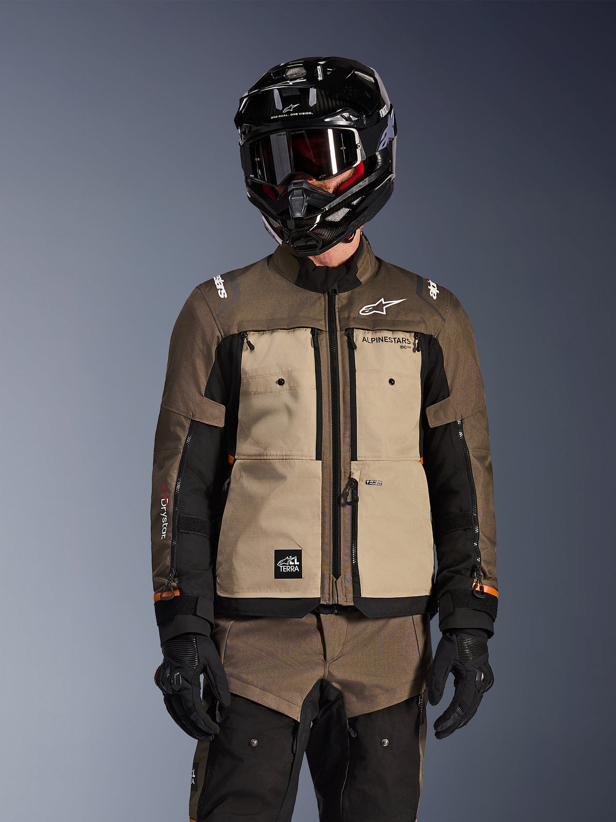 Motociclista che indossa la giacca Alpinestars Cusco Drystar in Walnut Aluminum Black, completo da moto adventure touring con casco nero, maschera e guanti neri, vista frontale che mostra la costruzione in tessuto tecnico, le tasche e il branding.