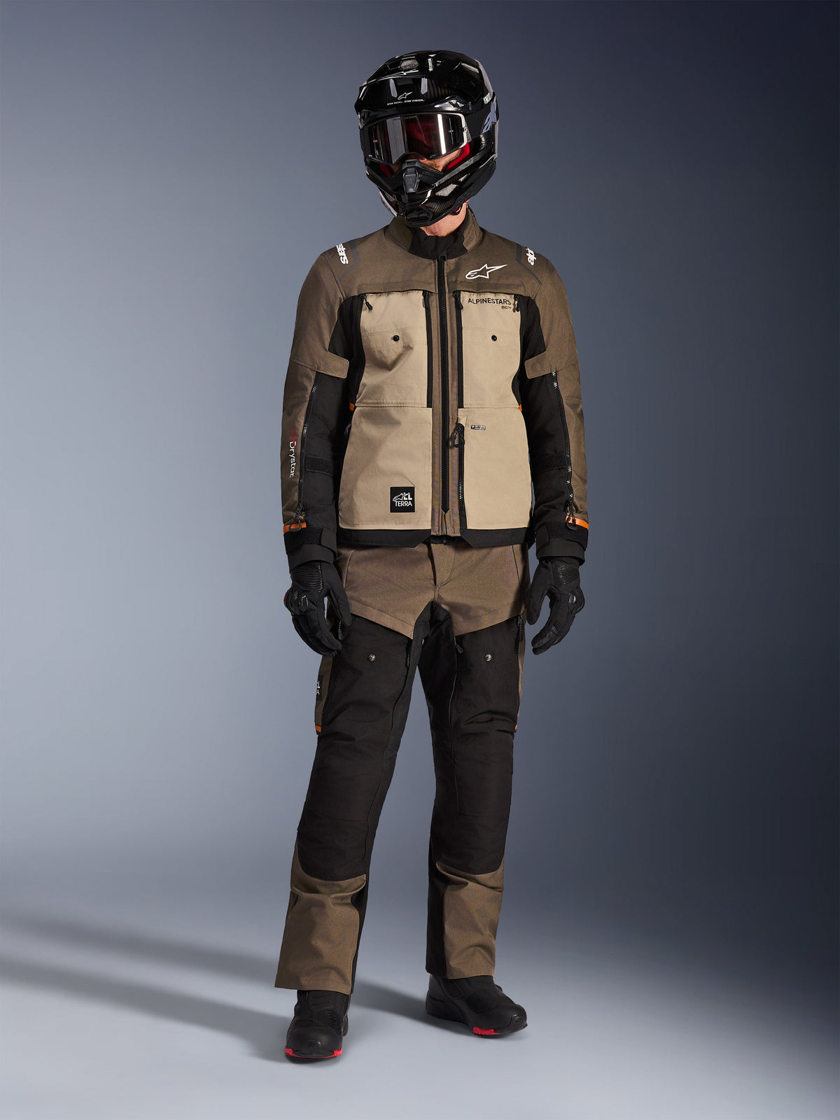Giacca Alpinestars Cusco Drystar®, colorazione Walnut Aluminum Black, giacca da moto adventure touring indossata con pantaloni coordinati e un casco nero, pannelli sul petto color sabbia e maniche in ripstop marrone, abbigliamento tecnico da guida per tutte le stagioni