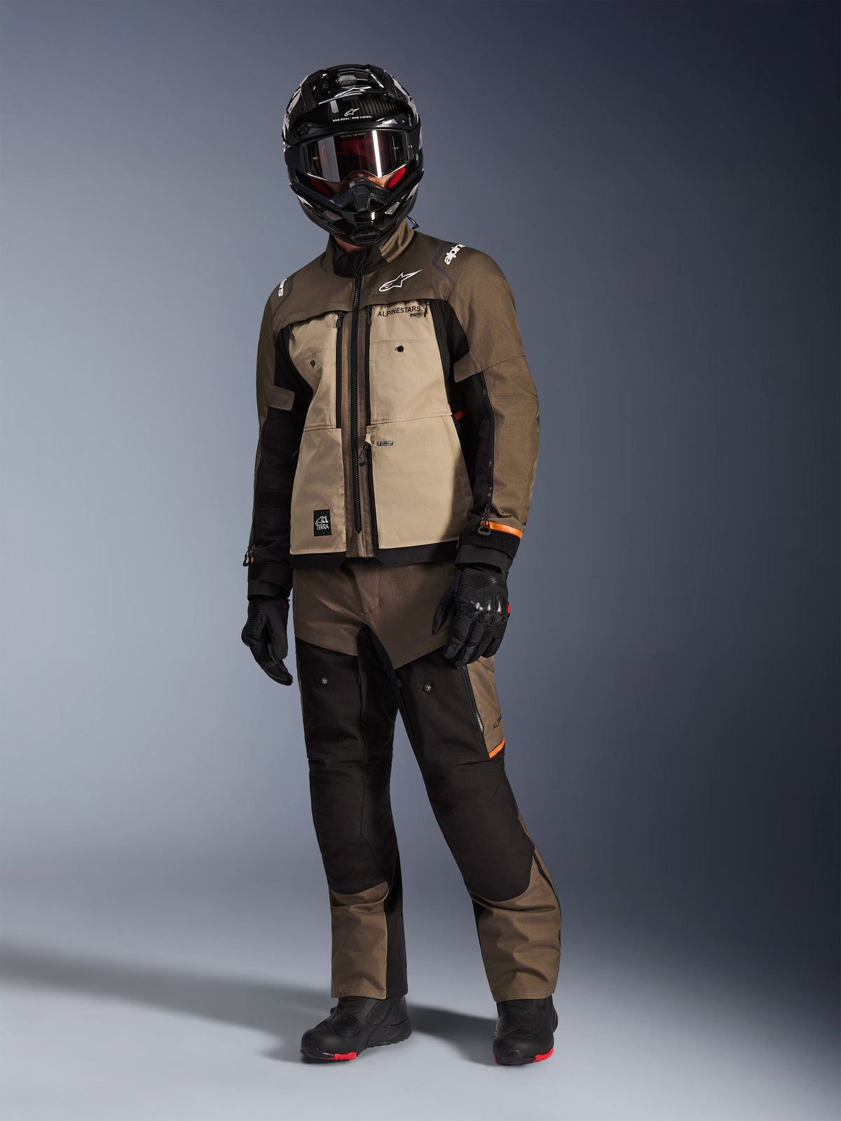 Giacca Alpinestars Cusco Drystar®, Walnut Aluminum Black, indossata da un pilota in abbigliamento completo da adventure touring, con pannelli sabbia e marroni e dettagli neri, coordinata con pantaloni abbinati, casco integrale nero con maschera e stivali neri, sfondo da studio.