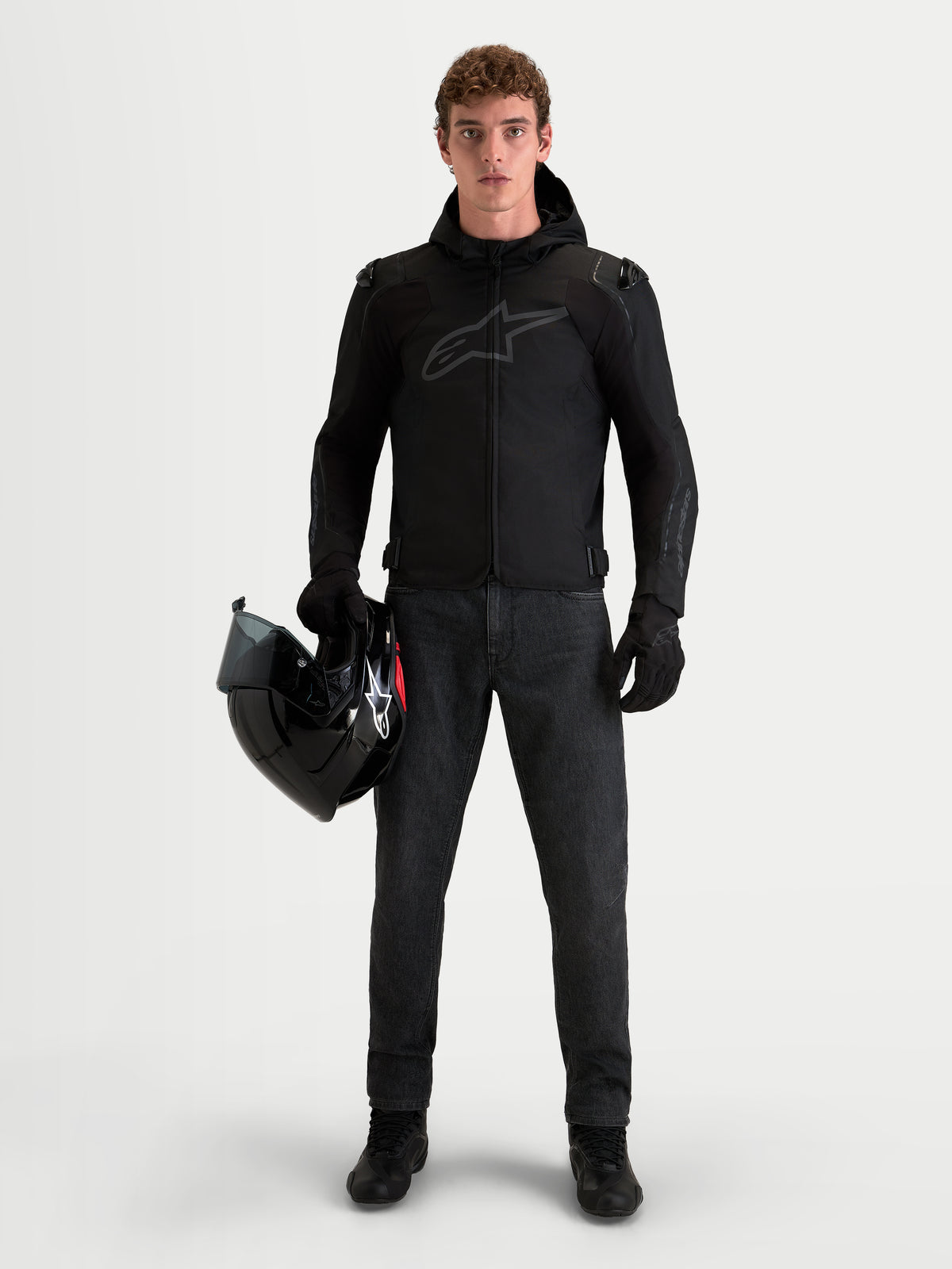 Alpinestars Zaca V 2 Waterproof Giacca, giacca da moto impermeabile in tessuto da uomo con cappuccio e protezione per le spalle, nero, indossata da un pilota che tiene in mano un casco