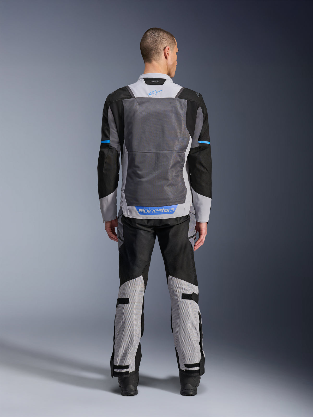 Giacca Alpinestars ST-2 Air, Ice Gray Dark Gray Black, vista posteriore su modello, con ampi pannelli di ventilazione in rete, dettagli blu e logo Alpinestars, abbinata a pantaloni da moto coordinati per adventure touring.