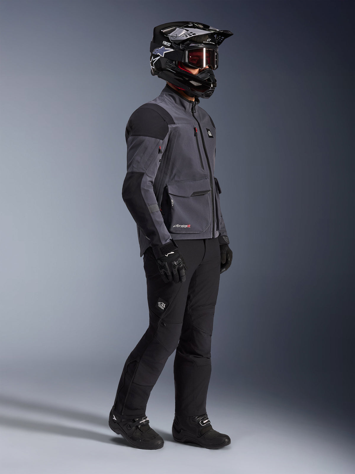 Motociclista che indossa la giacca Alpinestars Maxdura Drystar®XF Laminated Armored Shell in grigio scuro e nero, abbinata a pantaloni da avventura neri, casco nero con occhiali, guanti e stivali neri, scatto a figura intera che evidenzia l'equipaggiamento tecnico dual-sport.