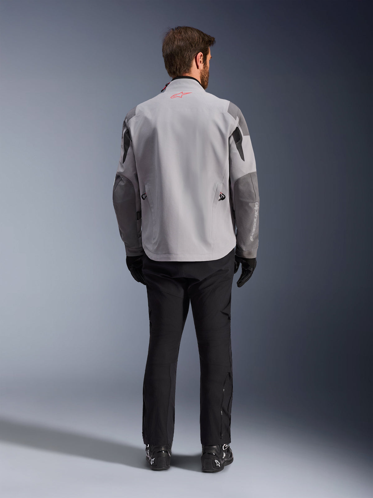 Alpinestars Maxdura Drystar®XF Laminated Armored Shell, giacca da moto grigio medio e grigio scuro, vista posteriore, caratterizzata da un corpo grigio chiaro con pannelli grigio scuro su spalle e maniche, piccolo logo a stella Alpinestars rosso sul colletto posteriore e marchio bianco sulla manica sinistra inferiore, abbinata a pantaloni e stivali da moto neri.