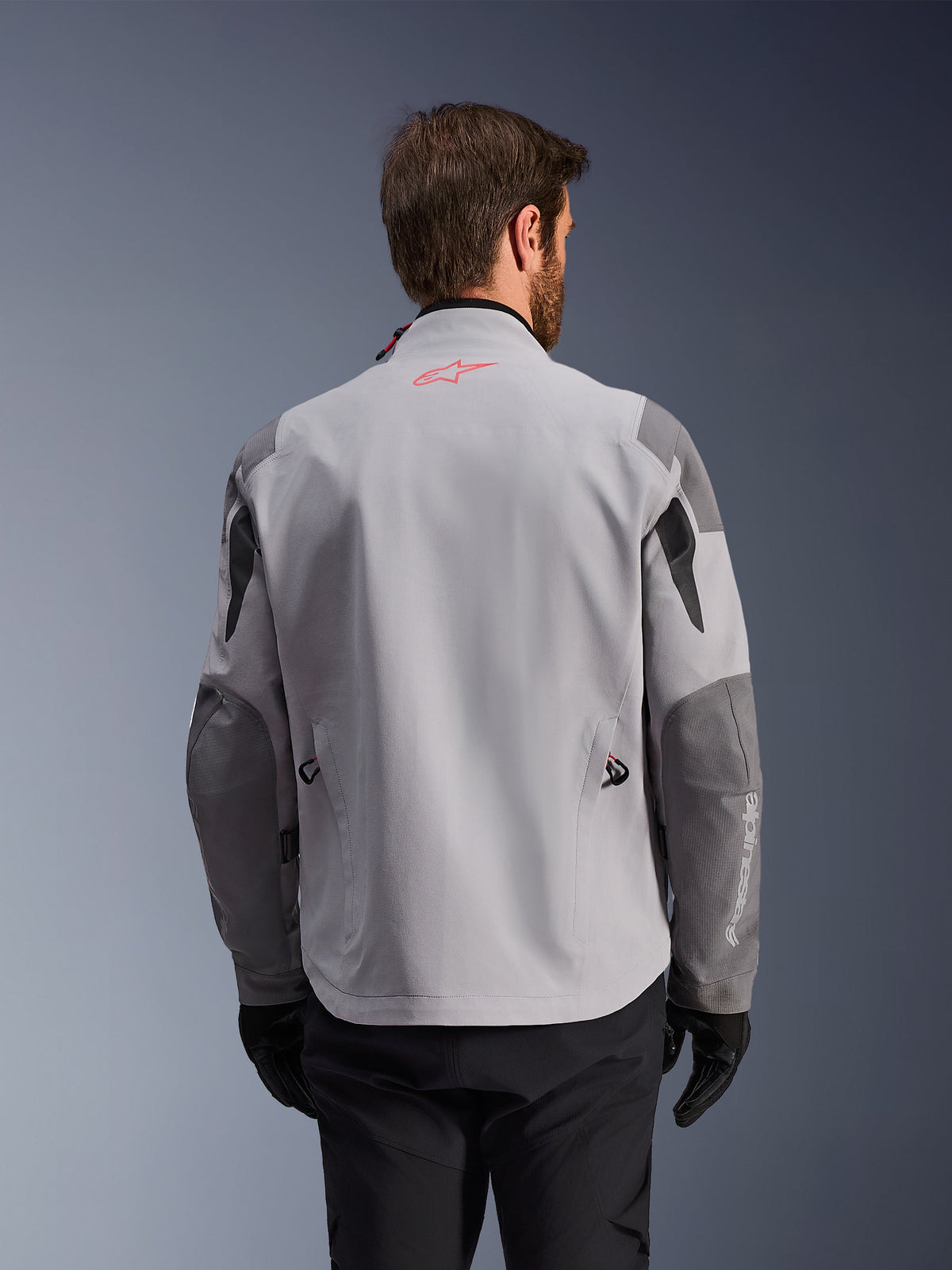 Alpinestars Maxdura Drystar®XF Laminated Armored Shell, vista posteriore di giacca da moto adventure, grigio medio e grigio scuro, con logo a stella rosso sul colletto e scritta verticale bianca sulla manica, indossata con guanti e pantaloni da guida neri