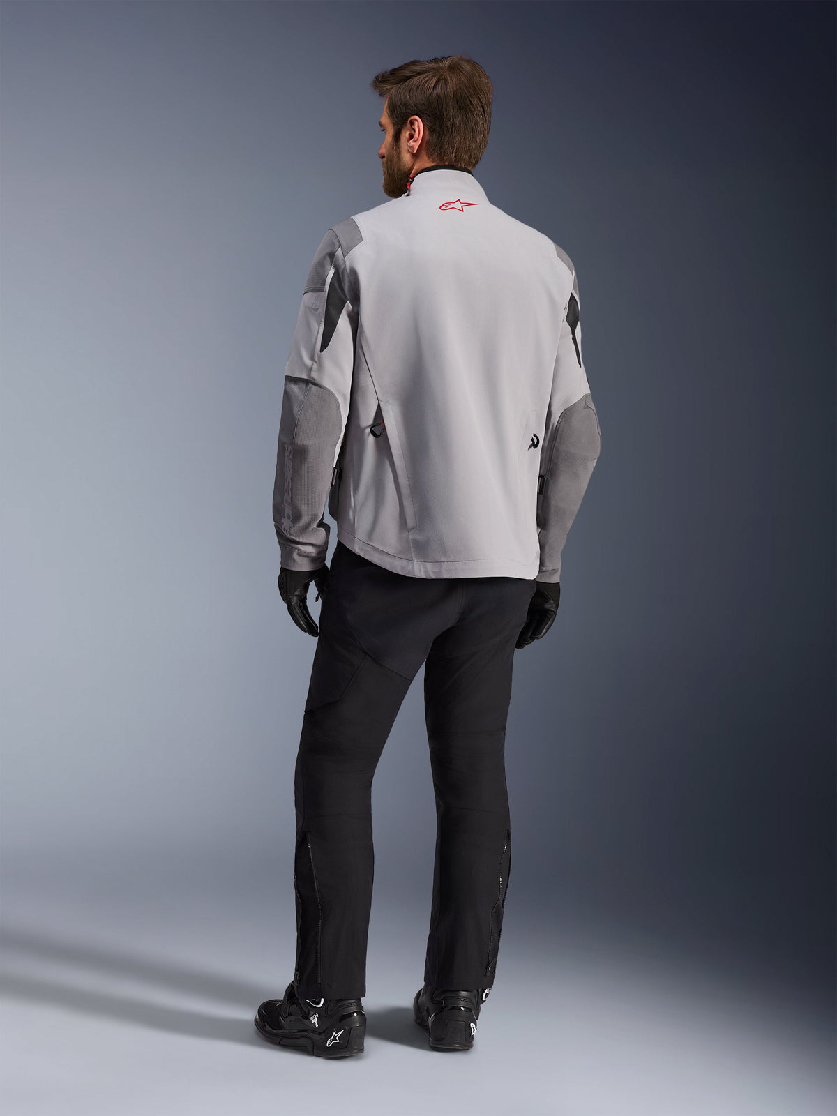 Maxdura Drystar®XF Laminated Armored Shell, giacca da moto, grigio medio e grigio scuro, vista posteriore con logo Alpinestars rosso sul colletto, rinforzi sulle maniche grigio scuro, indossata con pantaloni e stivali neri, abbigliamento per guida adventure