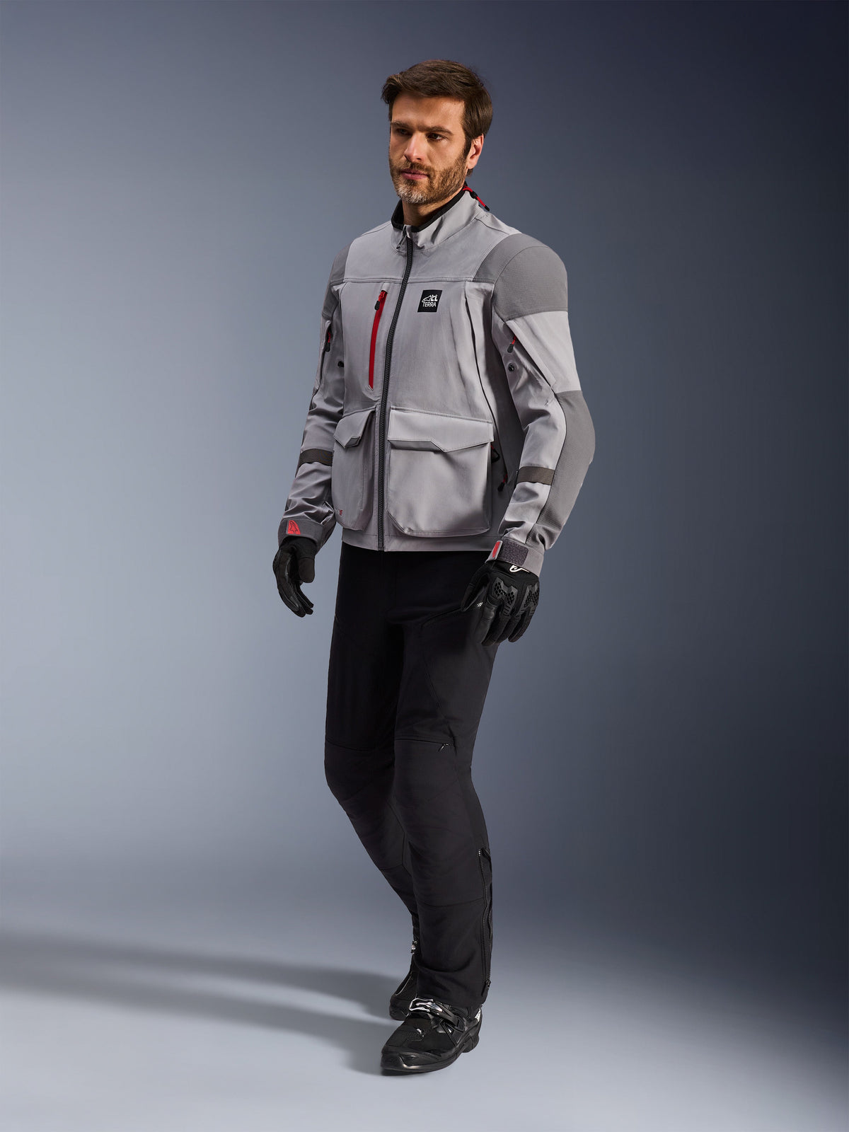 Alpinestars Maxdura Drystar®XF Laminated Armored Shell, giacca da moto adventure, grigio medio e grigio scuro, indossata da un modello con pantaloni da moto e guanti neri, con dettagli delle cerniere rossi e tasche cargo, abbigliamento tecnico professionale per adventure touring