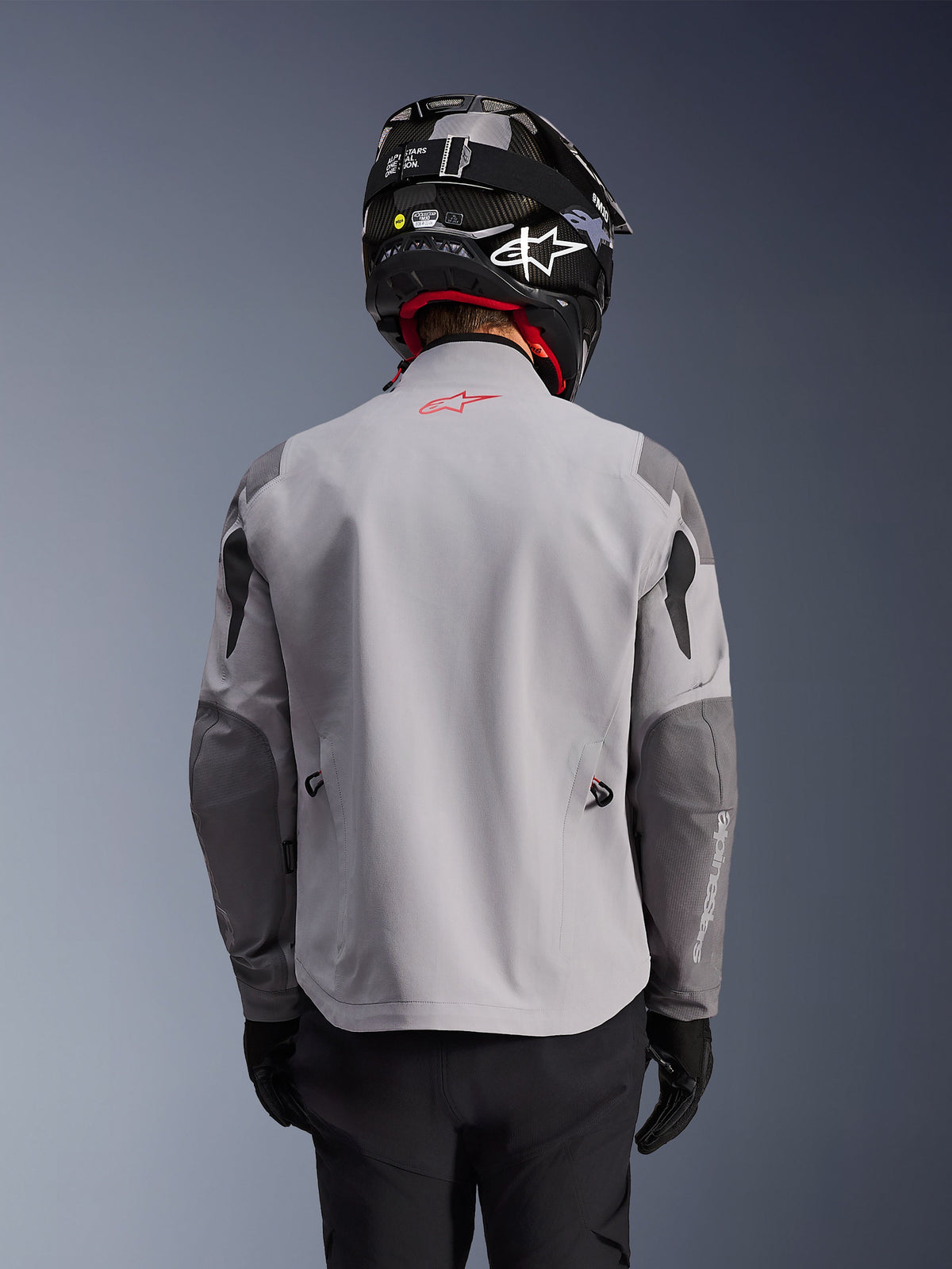 Alpinestars Maxdura Drystar®XF Laminated Armored Shell, giacca da moto adventure, grigio medio e grigio scuro, vista posteriore che mostra il corpo grigio chiaro con pannelli rinforzati grigio scuro su spalle e gomiti, logo Alpinestars rosso sulla parte superiore della schiena, indossata con casco e guanti neri