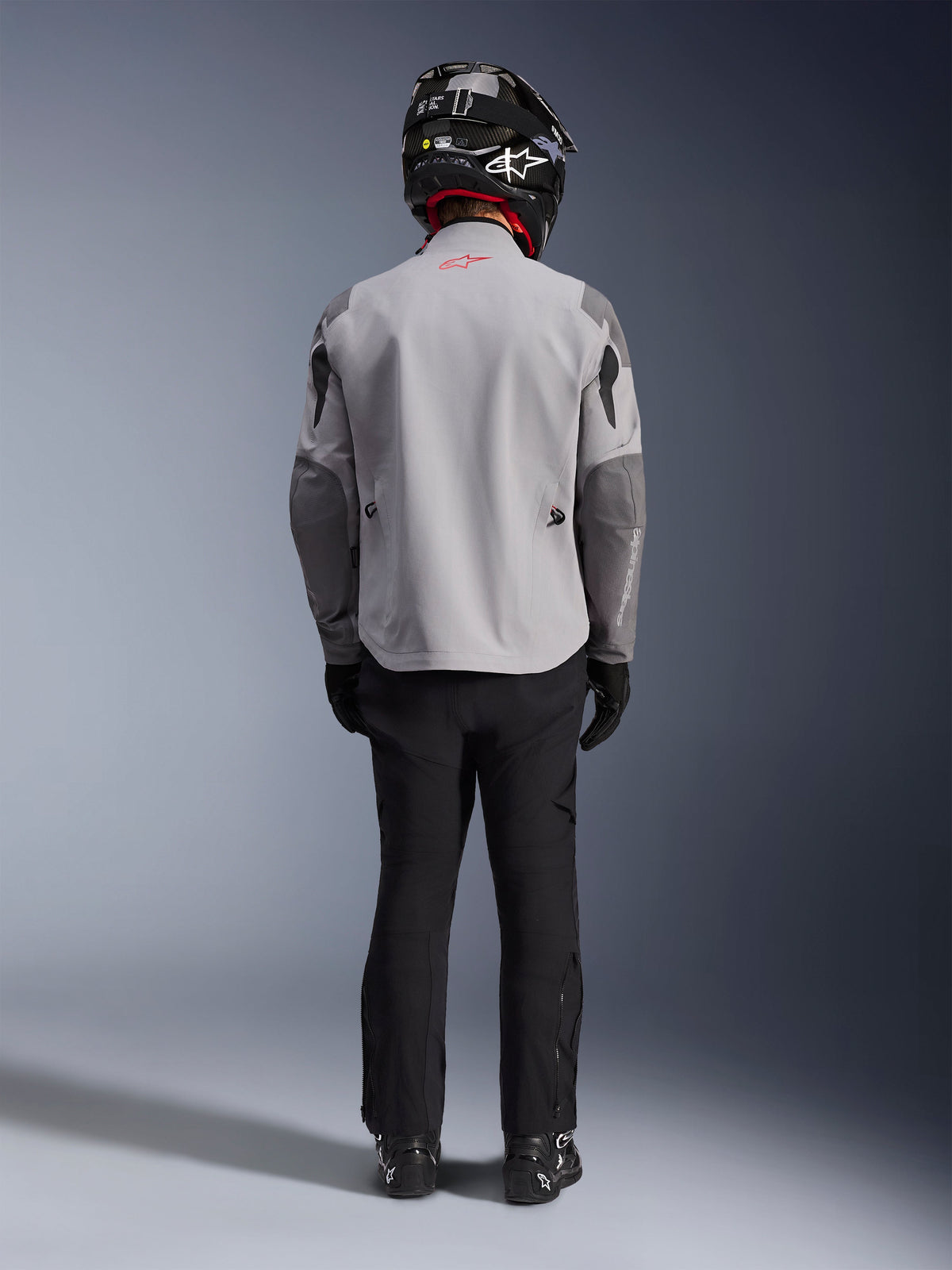 Alpinestars Maxdura Drystar®XF Laminated Armored Shell, giacca da moto grigio medio e grigio scuro, vista dal retro su un pilota, indossata con pantaloni neri, stivali neri e un casco bianco e nero, abbigliamento adventure touring
