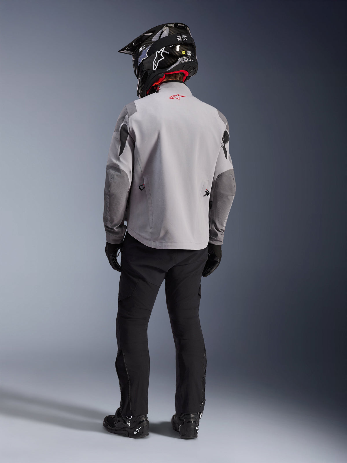 Vista posteriore di un motociclista con giacca Alpinestars Maxdura Drystar®XF Laminated Armored Shell in grigio medio e grigio scuro, logo con stella rossa sul colletto, pantaloni adventure neri, casco nero con trama in carbonio e stivali neri.