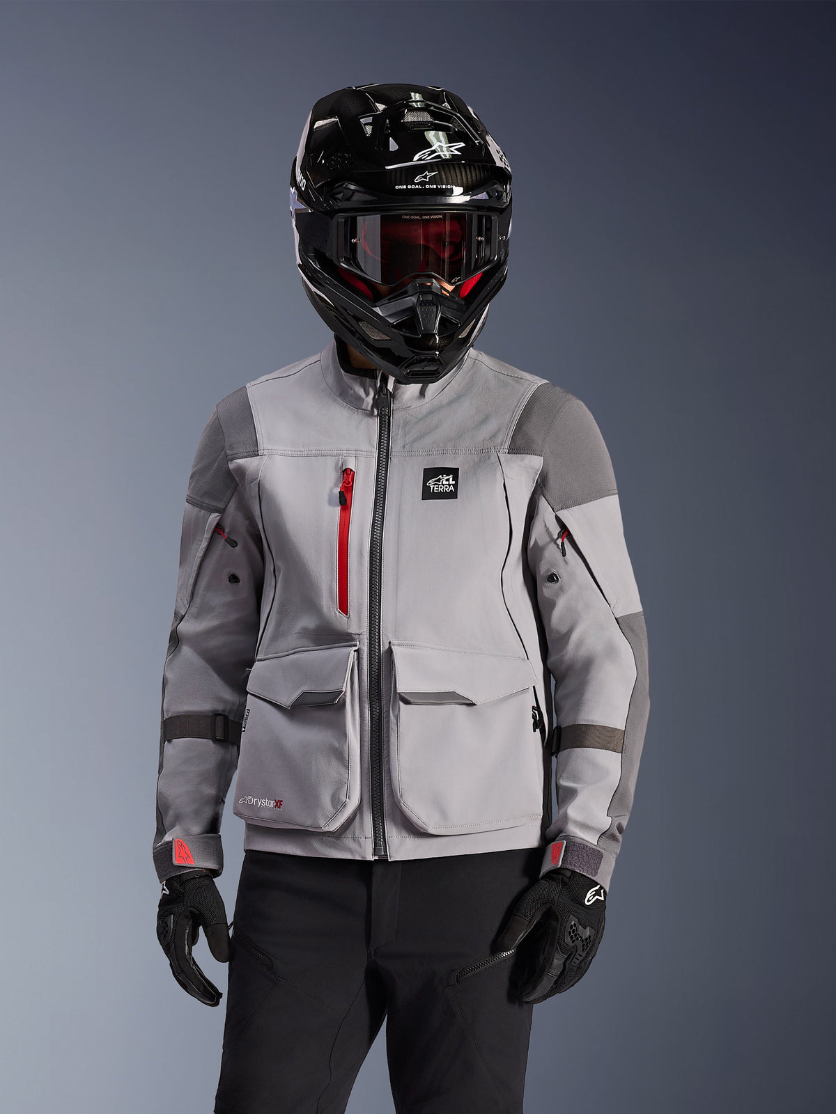 Alpinestars Maxdura Drystar®XF Laminated Armored Shell, giacca da moto adventure in grigio medio e grigio scuro, indossata con un casco nero con finitura in carbonio e occhiali con lenti rosse, con tasche cargo anteriori e una cerniera rossa sul petto, design tecnico per adventure touring.