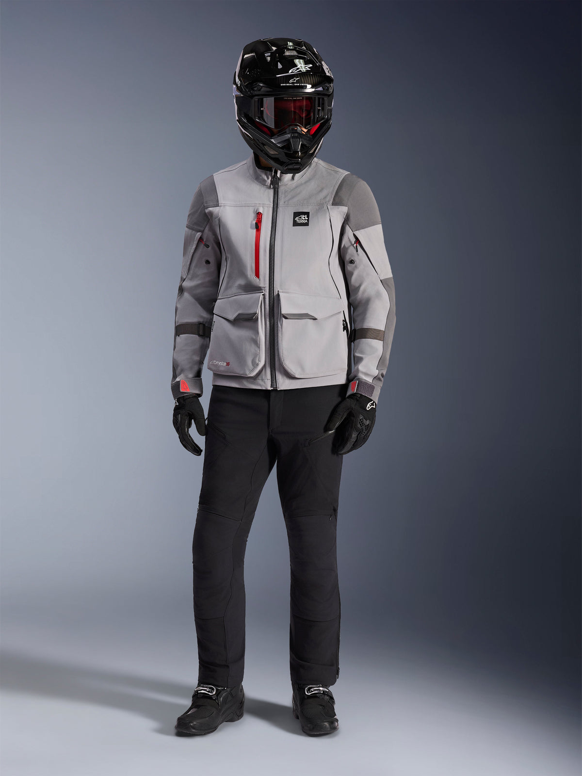 Giacca adventure Alpinestars Maxdura Drystar®XF Laminated Armored Shell, grigio medio e grigio scuro con dettagli rossi sulle cerniere, indossata da un pilota con casco nero, maschera rossa e pantaloni neri, abbigliamento professionale da moto per ogni condizione atmosferica