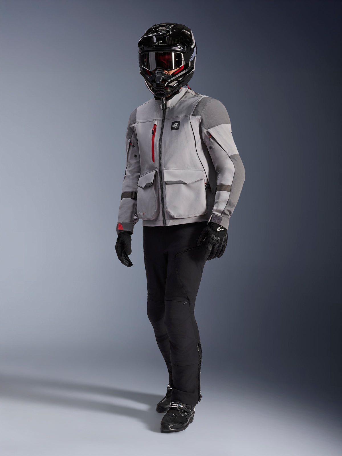 Pilota adventure che indossa Alpinestars Maxdura Drystar®XF Laminated Armored Shell, giacca da moto grigio medio e grigio scuro, casco adventure nero con maschera, pantaloni e guanti neri, con accenti rossi sulle cerniere e tasche cargo, abbigliamento tecnico per adventure touring