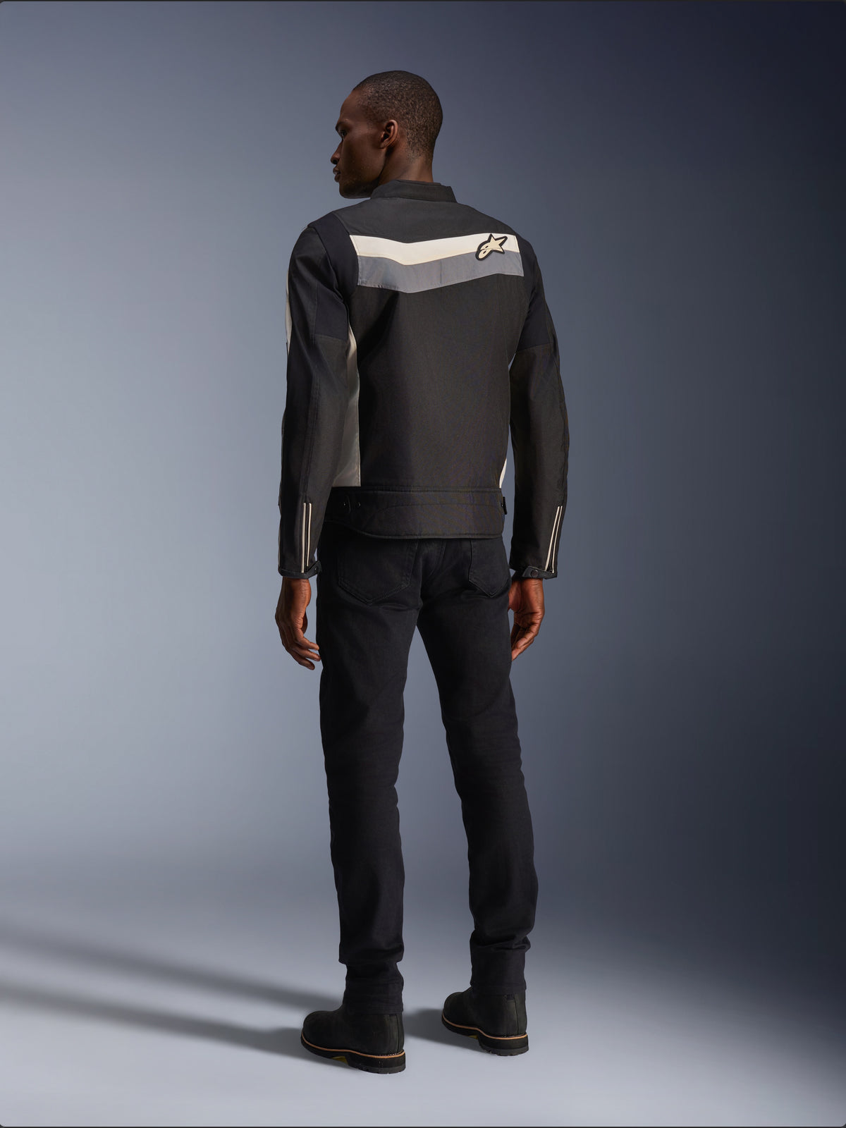 Modello maschile visto da dietro che indossa la giacca Alpinestars T-Dyno WR in Nero Grigio Scuro Ecru, giacca da moto in tessuto con striscia orizzontale e logo a stella sul retro, abbinata a pantaloni da guida e stivali neri, estetica urban motorcycle gear.