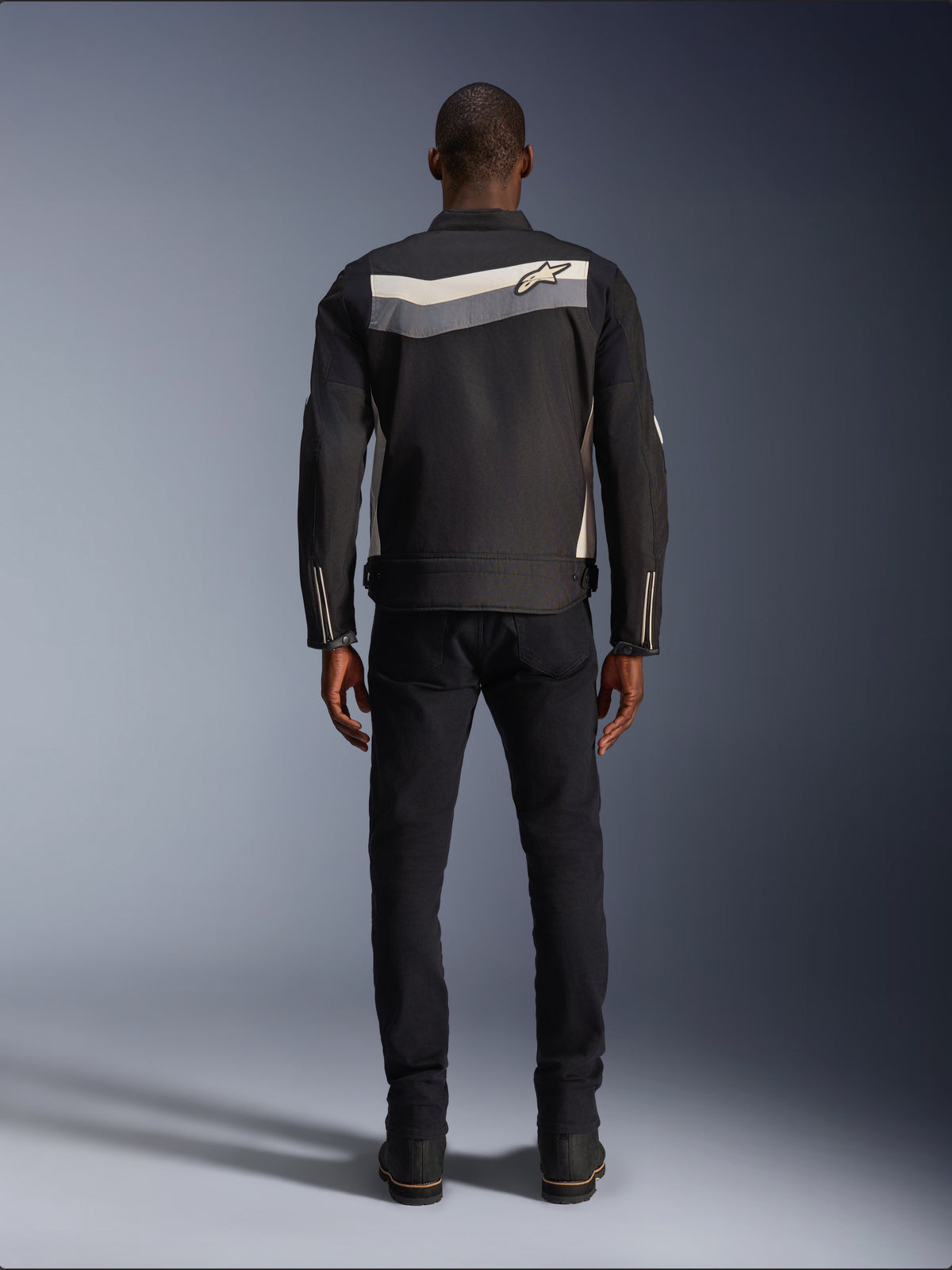 Giacca Alpinestars T-Dyno WR, giacca da moto in tessuto, nero, grigio scuro ed ecru, vista posteriore con striscia orizzontale sulle spalle e piccolo logo a stella, indossata da un modello con pantaloni e stivali neri, abbigliamento tecnico d'ispirazione vintage