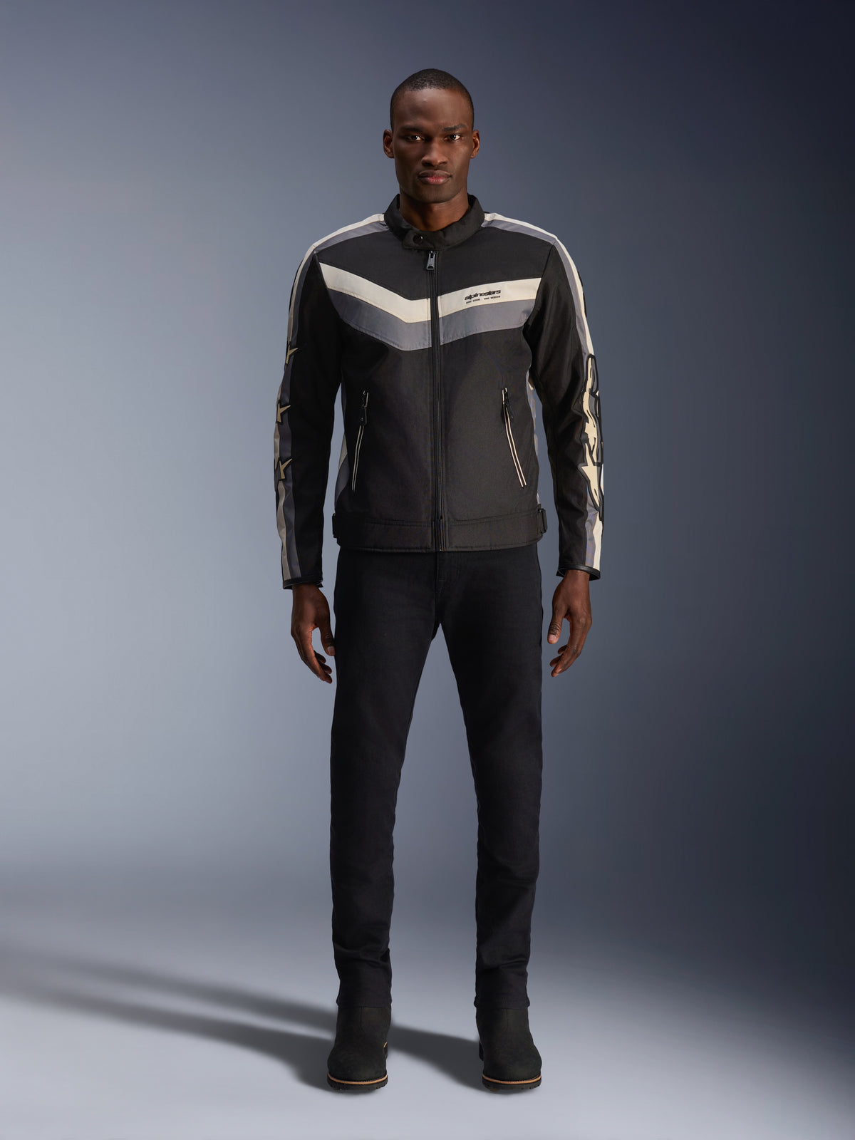 Giacca Alpinestars T-Dyno WR, nero, grigio scuro ed ecru, giacca da moto in tessuto con motivo chevron sul petto d'ispirazione vintage, indossata da un modello uomo con pantaloni e stivali da moto neri, abbigliamento stradale urban contemporary