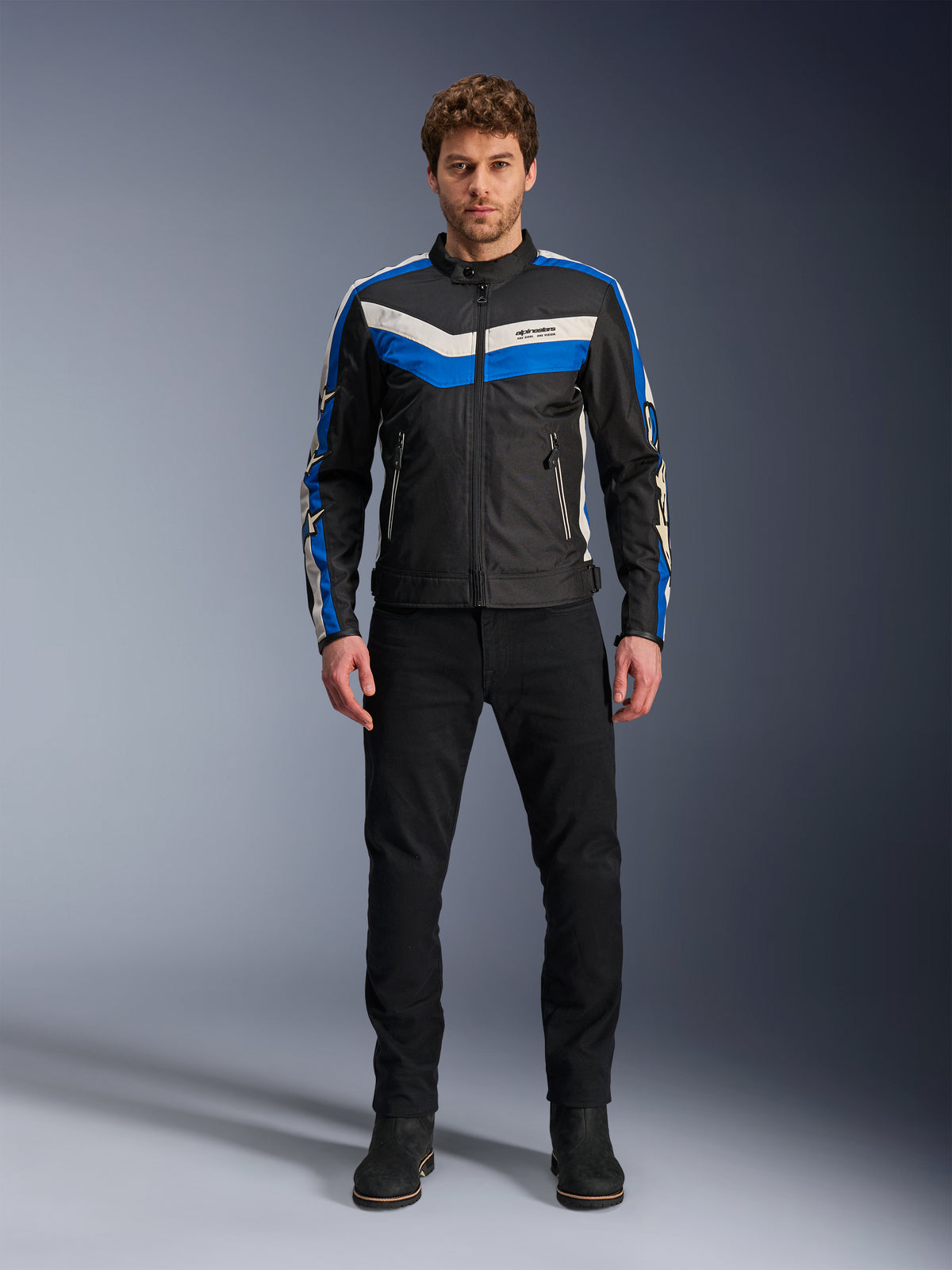 Giacca Alpinestars T-Dyno WR, giacca da moto in tessuto, nero, blu ed ecru, indossata da un modello con pantaloni e stivali neri, con strisce a chevron sul petto e design biker heritage