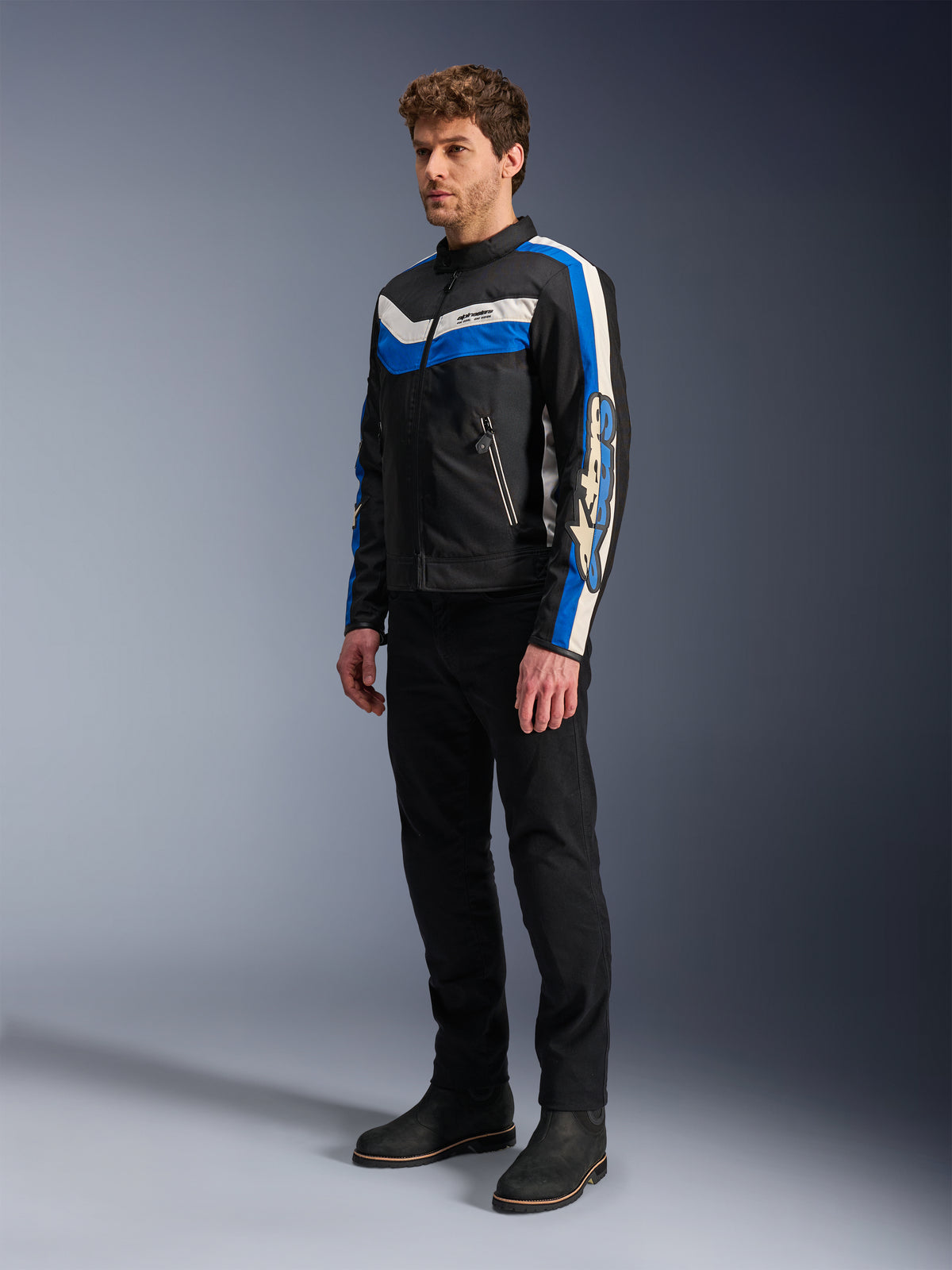 Alpinestars T-Dyno WR Jacket, nero, blu ed ecru, giacca da moto in tessuto indossata da un modello con pantaloni e stivali neri, design d'ispirazione vintage con vistose strisce blu ed ecru e loghi sulle maniche, fotografia in studio