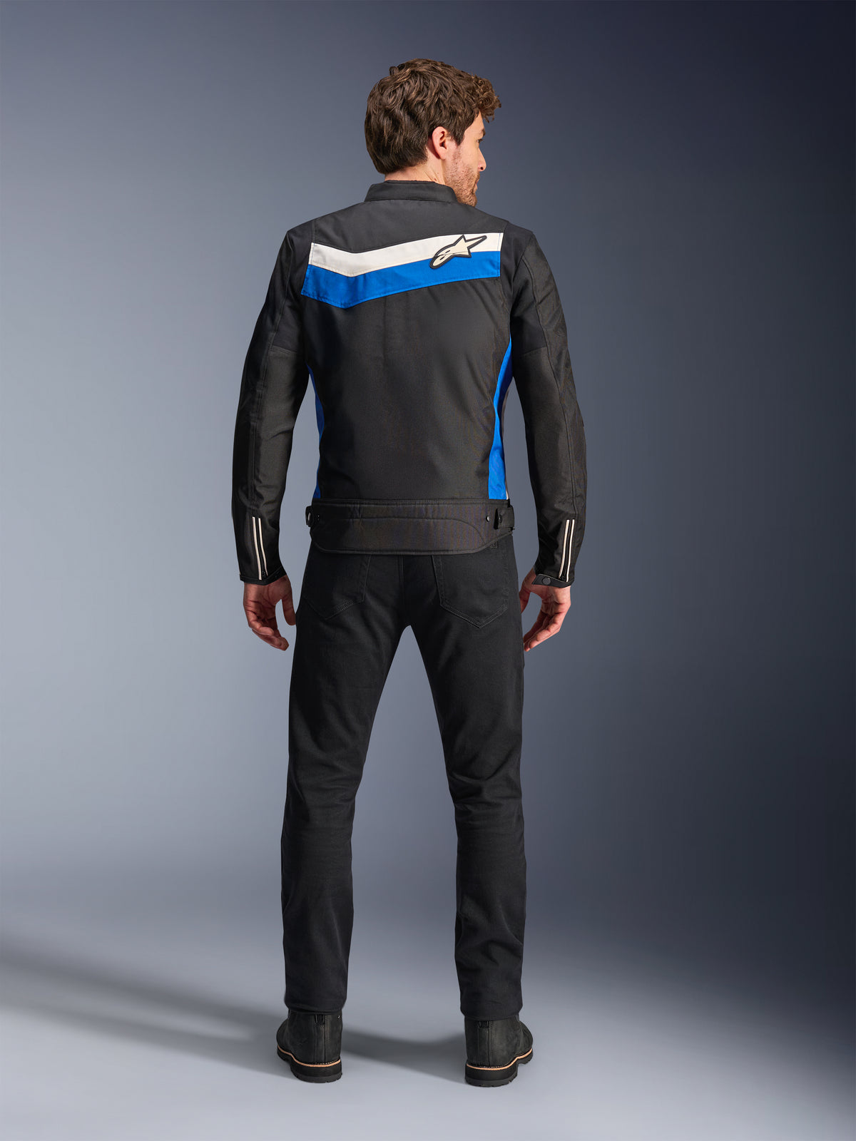Giacca Alpinestars T-Dyno WR, giacca da moto in tessuto, colorazione Nero Blu Ecru, vista posteriore completa su modello, caratterizzata da una striscia a chevron blu e ecru con logo Alpinestars, abbinata a pantaloni e stivali neri, abbigliamento da moto urbano d'ispirazione vintage