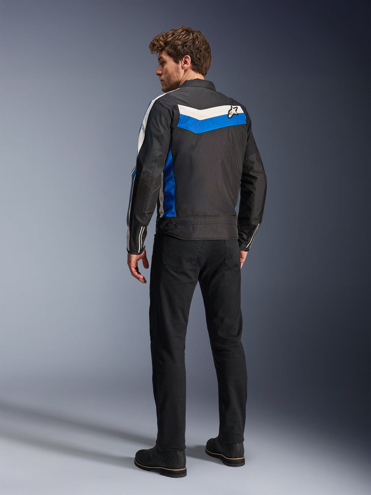 Giacca Alpinestars T-Dyno WR in Black Blue Ecru, vista posteriore su un modello maschile, caratterizzata da una striscia chevron bianca e blu sulle spalle con logo a stella, giacca da moto in tessuto d'ispirazione vintage, indossata con pantaloni e stivali neri