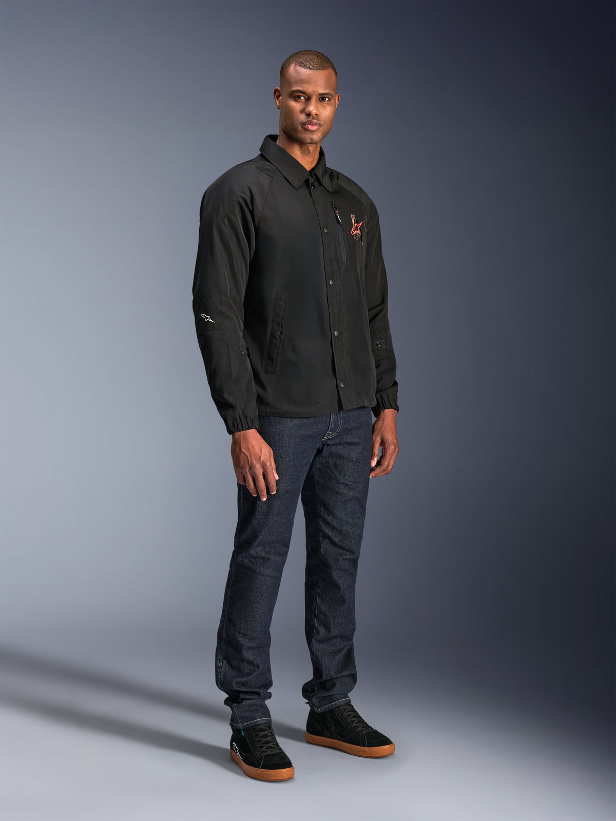 Giacca Alpinestars Revved WR in nero, giacca da moto in stile coach urbano indossata da un modello con jeans blu scuro e sneakers nere, con chiusura frontale a bottoni a pressione e logo ricamato, design minimalista e sportivo per la guida su strada