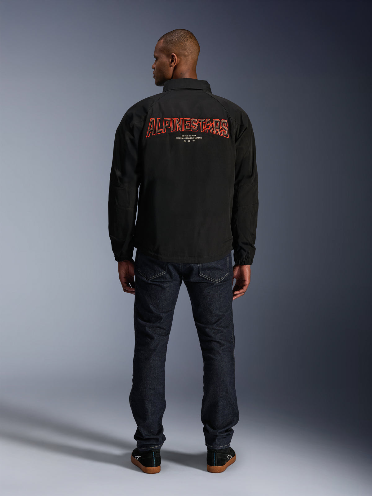 Alpinestars Giacca Revved WR, nera, vista posteriore con grande ricamo rosso 'ALPINESTARS' effetto 3D, giacca stile urban coach, indossata con jeans denim scuro e sneakers nere, abbigliamento da moto minimalista