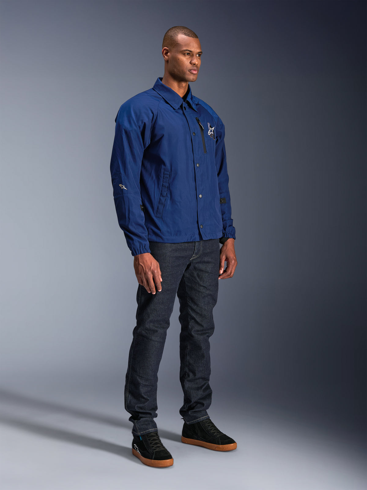 Giacca Alpinestars Revved WR, giacca da moto blu, indossata da un modello uomo con jeans in denim scuro e sneakers nere, caratterizzata da una silhouette da giacca da allenatore resistente all'acqua con chiusura frontale a bottoni a pressione e logo minimalista sul petto.