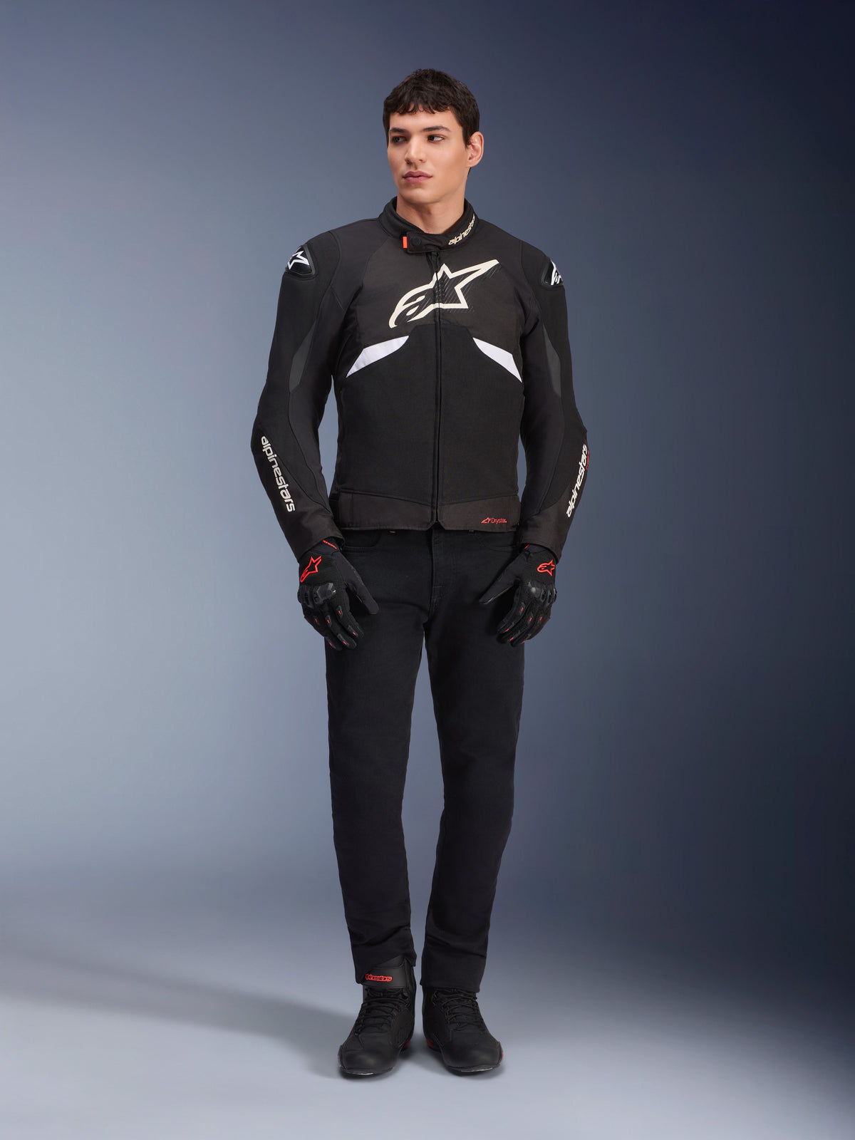 Modello che indossa la giacca Alpinestars T-GP R V3 Drystar® in nero e bianco, giacca sportiva in tessuto impermeabile con grande logo Alpinestars bianco sul petto, indossata con pantaloni e guanti da moto neri, ritratto professionale in studio che mette in risalto la silhouette sportiva.