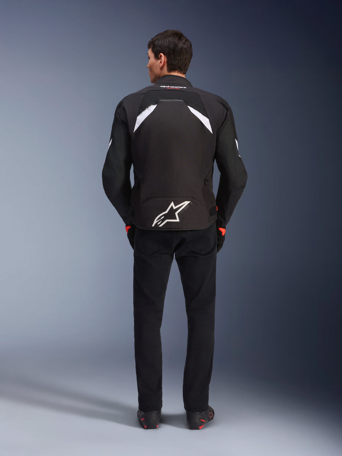 Giacca Alpinestars T-GP R V3 Drystar®, nero e bianco, vista posteriore, giacca da moto in tessuto impermeabile con un grande logo a stella bianco sulla parte inferiore della schiena e dettagli bianchi sulle spalle, abbinata a pantaloni e stivali neri, design sportivo derivato dalle corse.