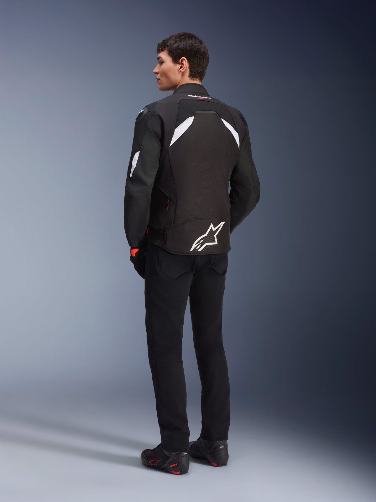 Modello maschile visto di spalle che indossa la giacca Alpinestars T-GP R V3 Drystar® in nero e bianco, con dettagli bianchi sulle spalle e un grande logo bianco sulla parte bassa della schiena, abbinata a pantaloni e stivali da moto neri, giacca da moto sportiva in tessuto.