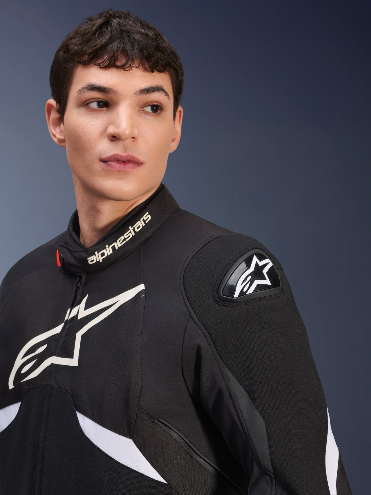 Giacca Alpinestars T-GP R V3 Drystar®, nero e bianco, indossata da un modello, con loghi Alpinestars bianchi sul colletto e sul petto, slider per spalle DFS nero con stella bianca, scatto lifestyle dettagliato di una giacca sportiva in tessuto impermeabile
