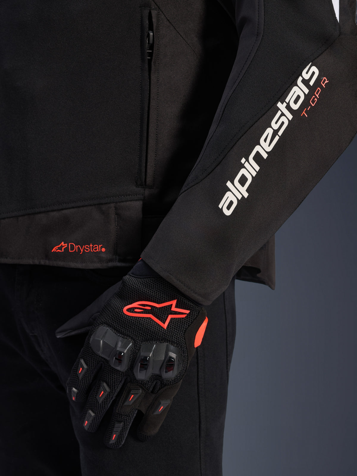 Giacca Alpinestars T-GP R V3 Drystar®, giacca da moto in tessuto nero, vista ravvicinata che mostra il marchio Alpinestars bianco e T-GP R arancione sulla manica, logo Drystar arancione sull'orlo, abbinata a guanti protettivi in mesh nero con accenti rossi.