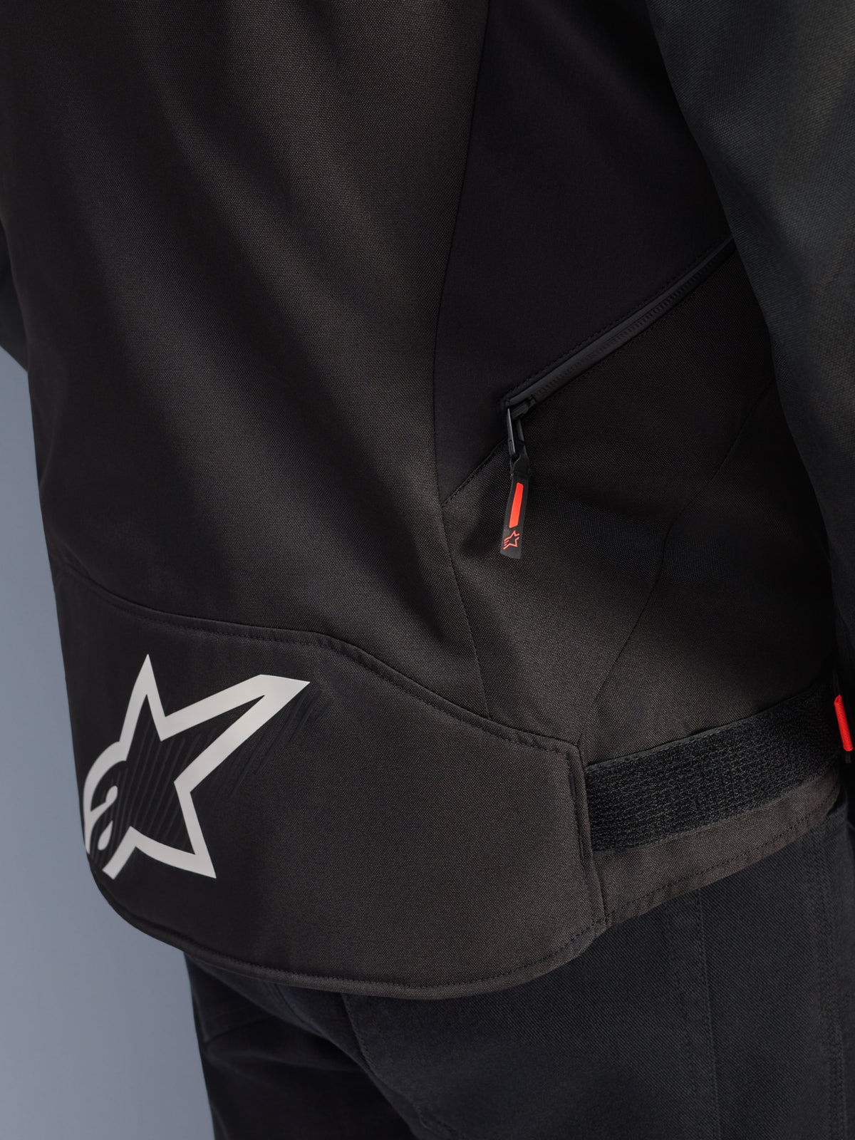 Giacca Alpinestars T-GP R V3 Drystar®, nero e bianco, vista ravvicinata della parte inferiore posteriore con un grande logo a stella Alpinestars bianco, una tasca laterale con cerniera e linguetta rossa e un cinturino di regolazione in vita in velcro, materiale tessile resistente.