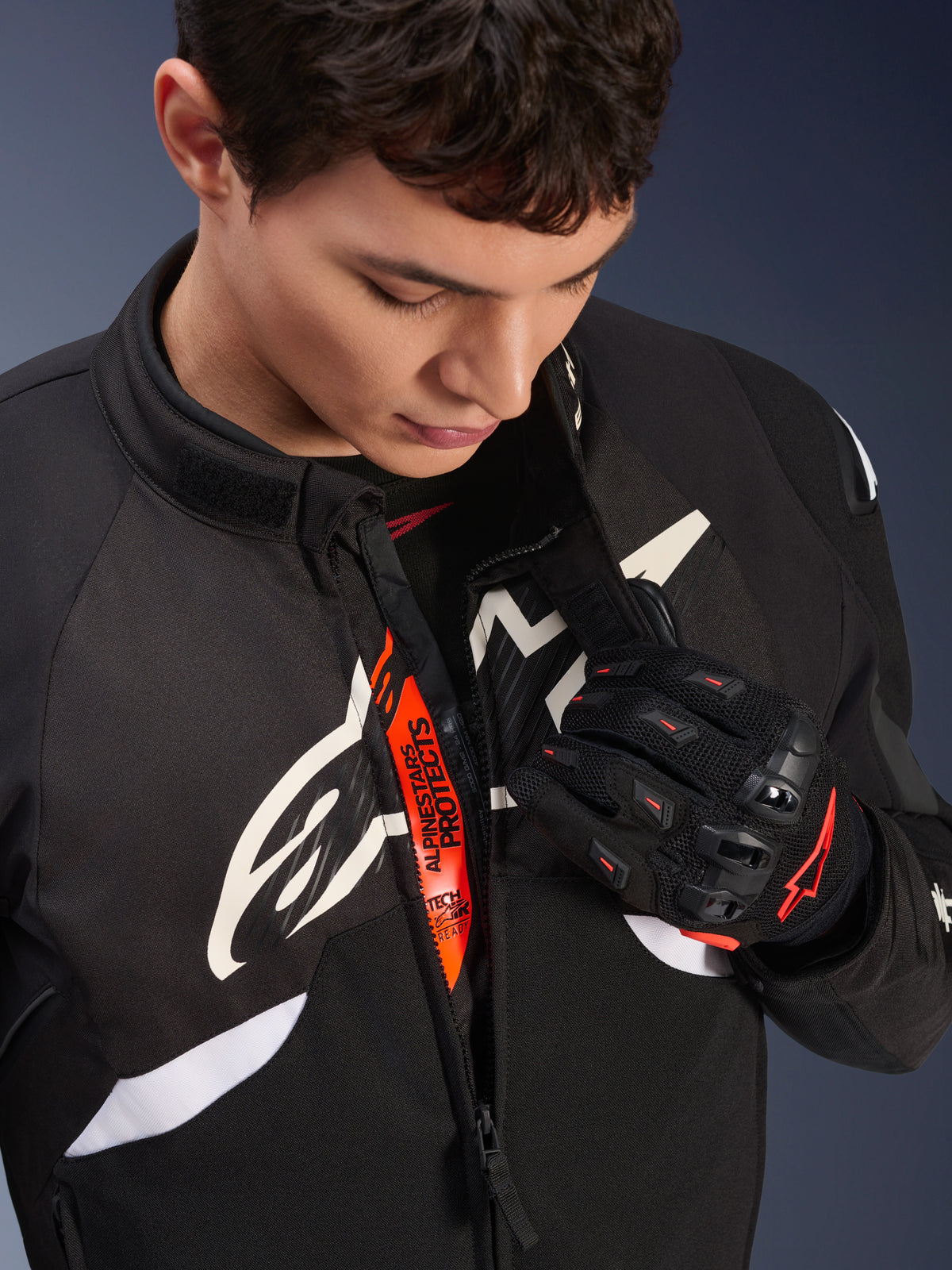 Giacca Alpinestars T-GP R V3 Drystar®, giacca sportiva da moto in tessuto nero e bianco, parzialmente aperta per rivelare la fascia interna arancione 'Alpinestars Protects', indossata con guanti in mesh neri e rossi, abbigliamento protettivo di derivazione racing.