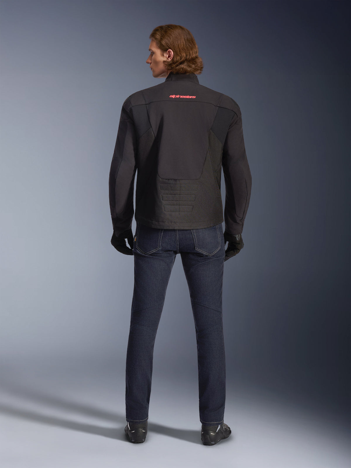 Giacca Alpinestars SMX Waterproof, nero, vista posteriore indossata da un modello, con logo rosso sulla parte superiore della schiena, orlo posteriore allungato per una maggiore copertura durante la guida, abbinata a jeans da moto blu scuro e guanti neri.