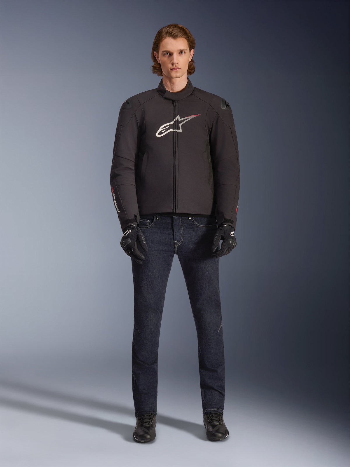 Modello che indossa la giacca Alpinestars SMX Waterproof in nero con logo bianco e rosso, abbinata a pantaloni da moto in denim scuro, guanti neri e stivali da guida, outfit sportivo da moto per tutte le condizioni atmosferiche.