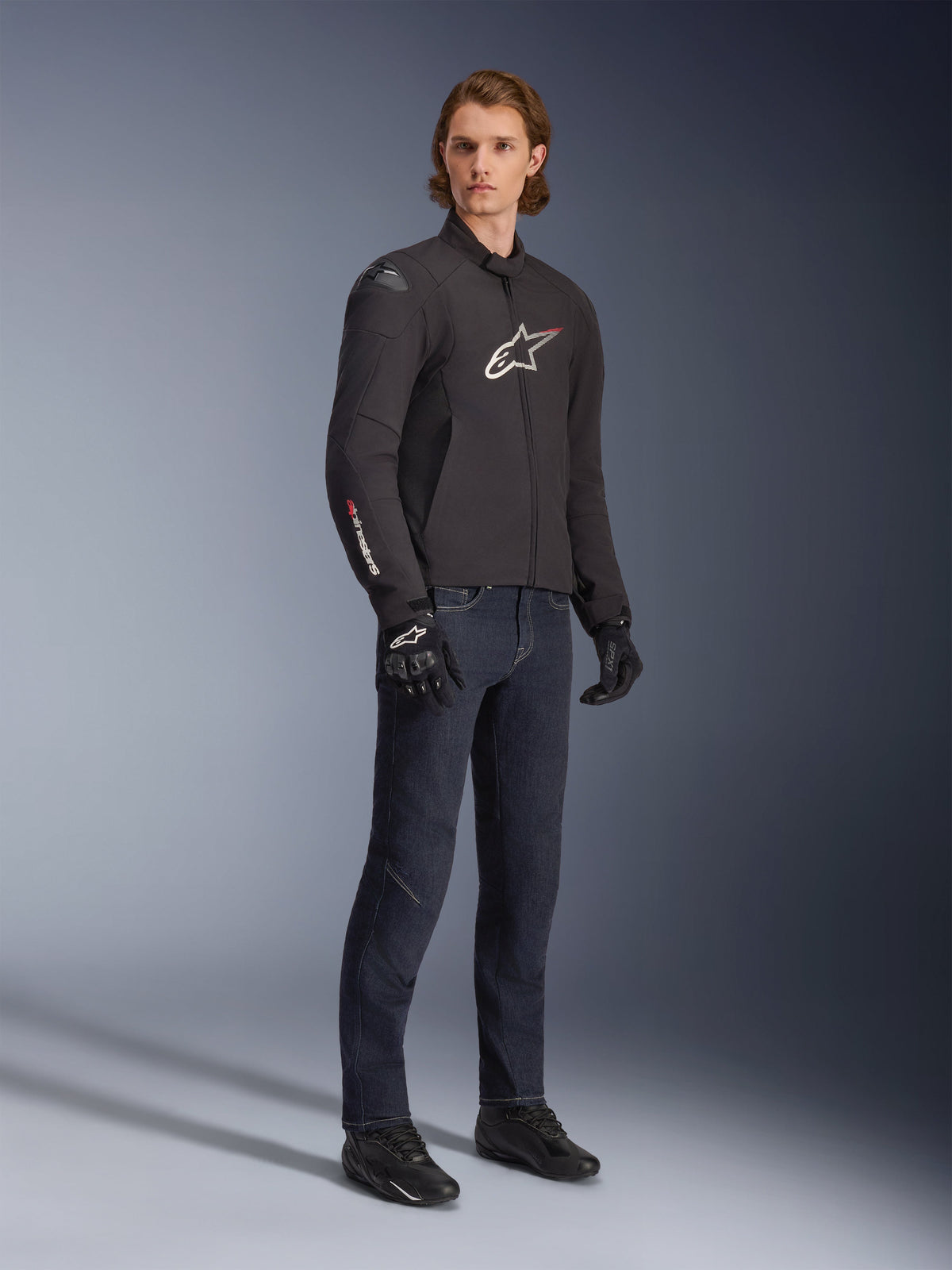 Modello che indossa la giacca Alpinestars SMX Waterproof, nera con accenti del logo bianchi e rossi, abbinata a pantaloni in denim blu scuro e guanti da moto neri, elegante giacca da moto in tessuto con vestibilità sportiva.