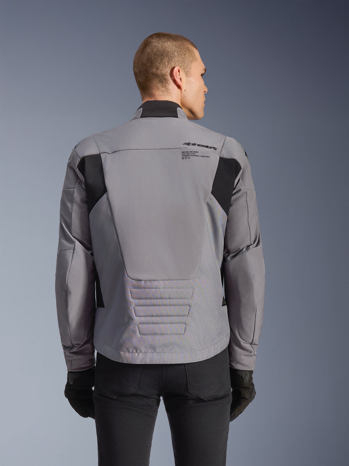 Giacca impermeabile Alpinestars SMX, grigio con accenti neri, vista posteriore che mostra il logo sulla schiena e il profilo posteriore allungato, indossata da un modello con pantaloni e guanti neri