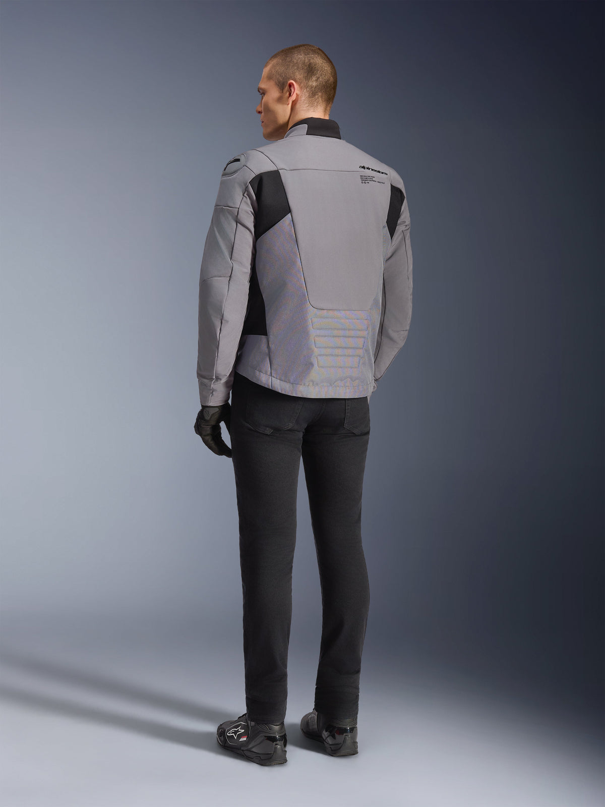 Giacca Alpinestars SMX Waterproof in grigio scuro e nero, vista posteriore su un modello, indossata con pantaloni e stivali neri, caratterizzata da costruzione in tessuto tecnico con inserti neri e logo Alpinestars sulla spalla.