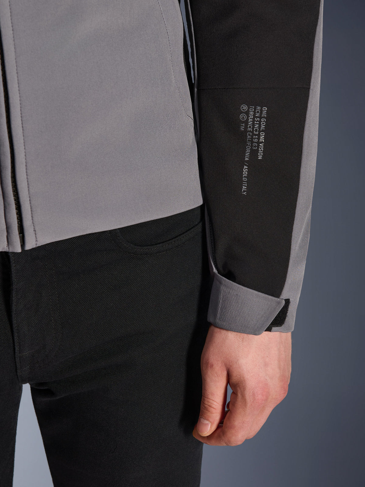 Giacca Alpinestars SMX Waterproof, grigio scuro e nero, primo piano della manica con testo stampato bianco e dettagli sulla storia del marchio, polsino regolabile in velcro, design sportivo in tessuto per la guida in moto in ogni condizione atmosferica