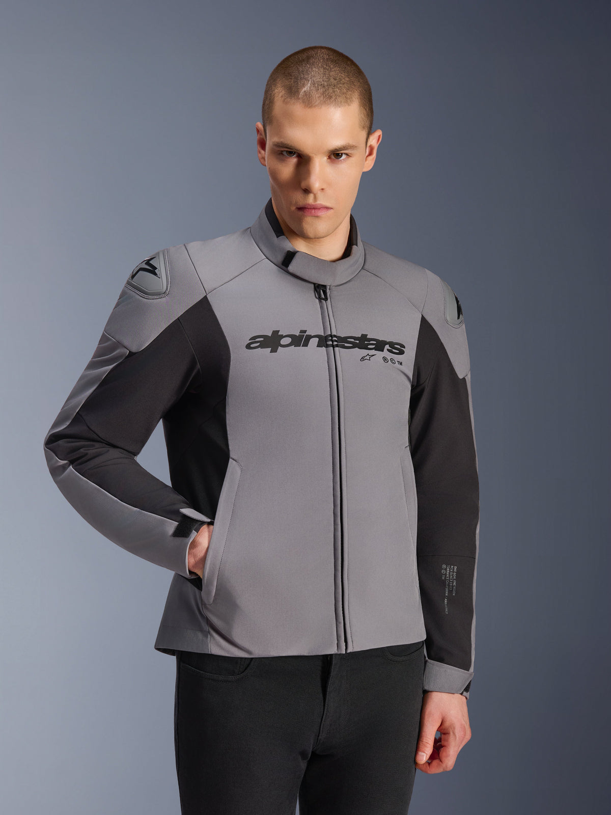 Giacca impermeabile Alpinestars SMX, grigio scuro e nero, indossata da un modello uomo, con logo 'alpinestars' sul petto, protezione per le spalle DFS Lite, design sportivo in tessuto e softshell per guida su strada