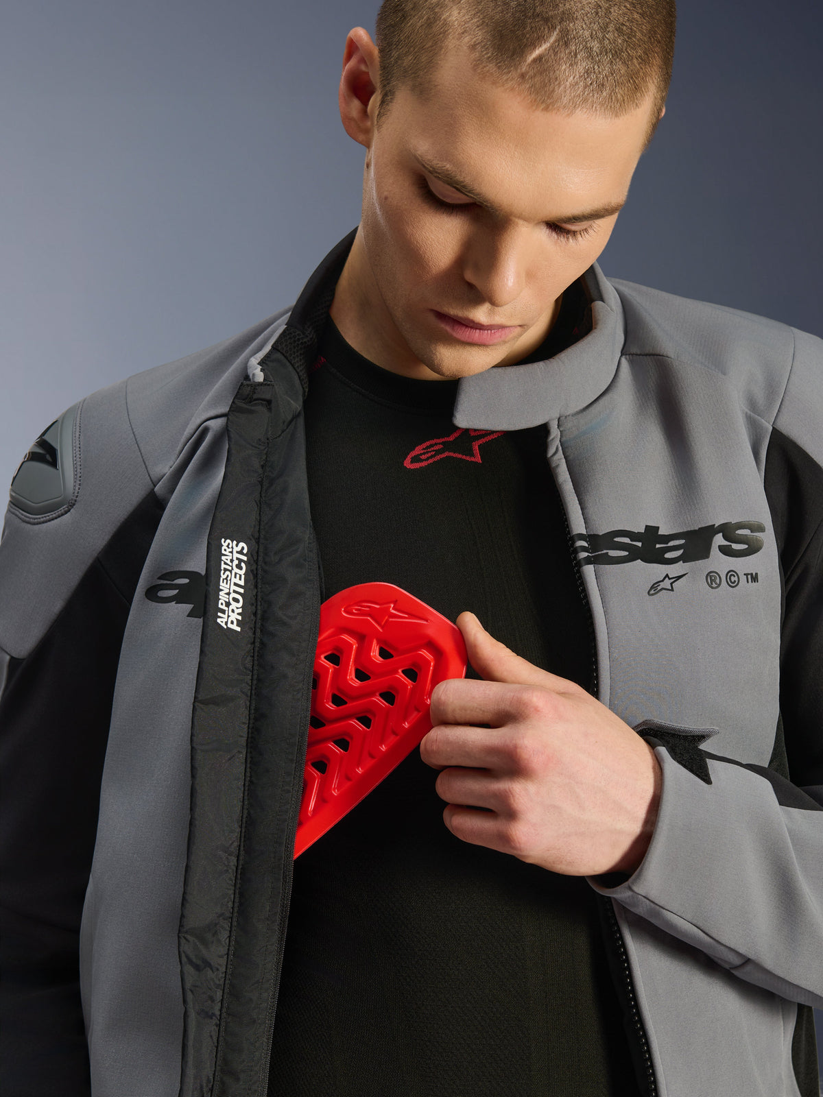Modello che indossa la giacca Alpinestars SMX Waterproof in grigio scuro e nero, mentre inserisce una protezione toracica Nucleon rossa in una tasca interna, mostrando la compatibilità della giacca con le protezioni e le sue caratteristiche protettive.