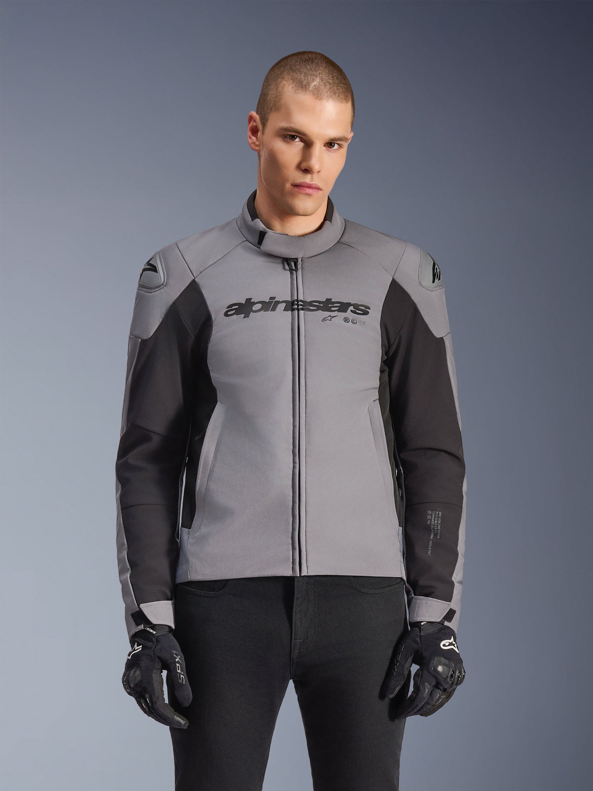 Giacca Alpinestars SMX Waterproof, giacca moto sportiva da strada, grigio scuro e nero, con colletto alto e slider sulle spalle DFS Lite, indossata con guanti SMX neri, vestibilità atletica per la guida in ogni condizione atmosferica