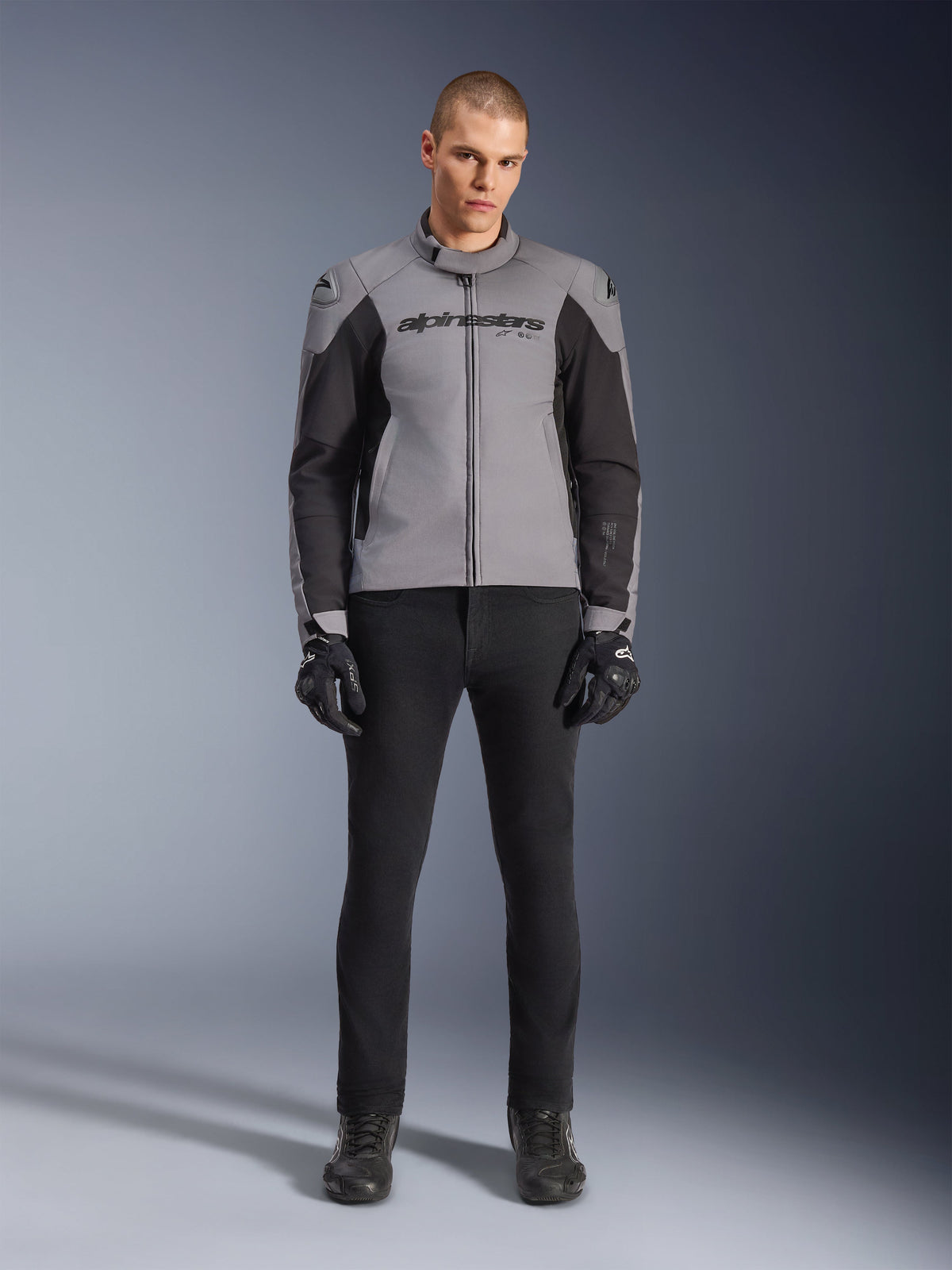 Giacca Alpinestars SMX Waterproof, grigio scuro e nero, vista frontale indossata da un modello, giacca da moto sportiva in tessuto con pannelli laterali neri e marchio Alpinestars sul petto, abbinata a pantaloni da guida e guanti neri.