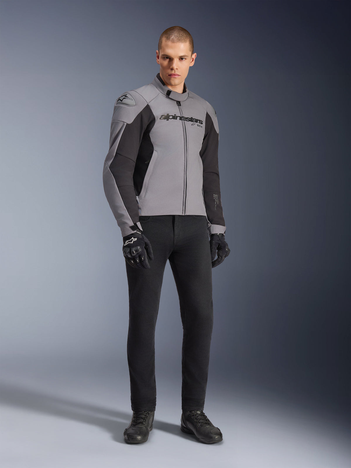 Giacca Alpinestars SMX Waterproof, giacca da moto, grigio scuro e nero, indossata da un modello con guanti da guida neri e pantaloni neri, con logo evidente sul petto e design tecnico con mappatura sportiva per la guida su strada in ogni condizione atmosferica.