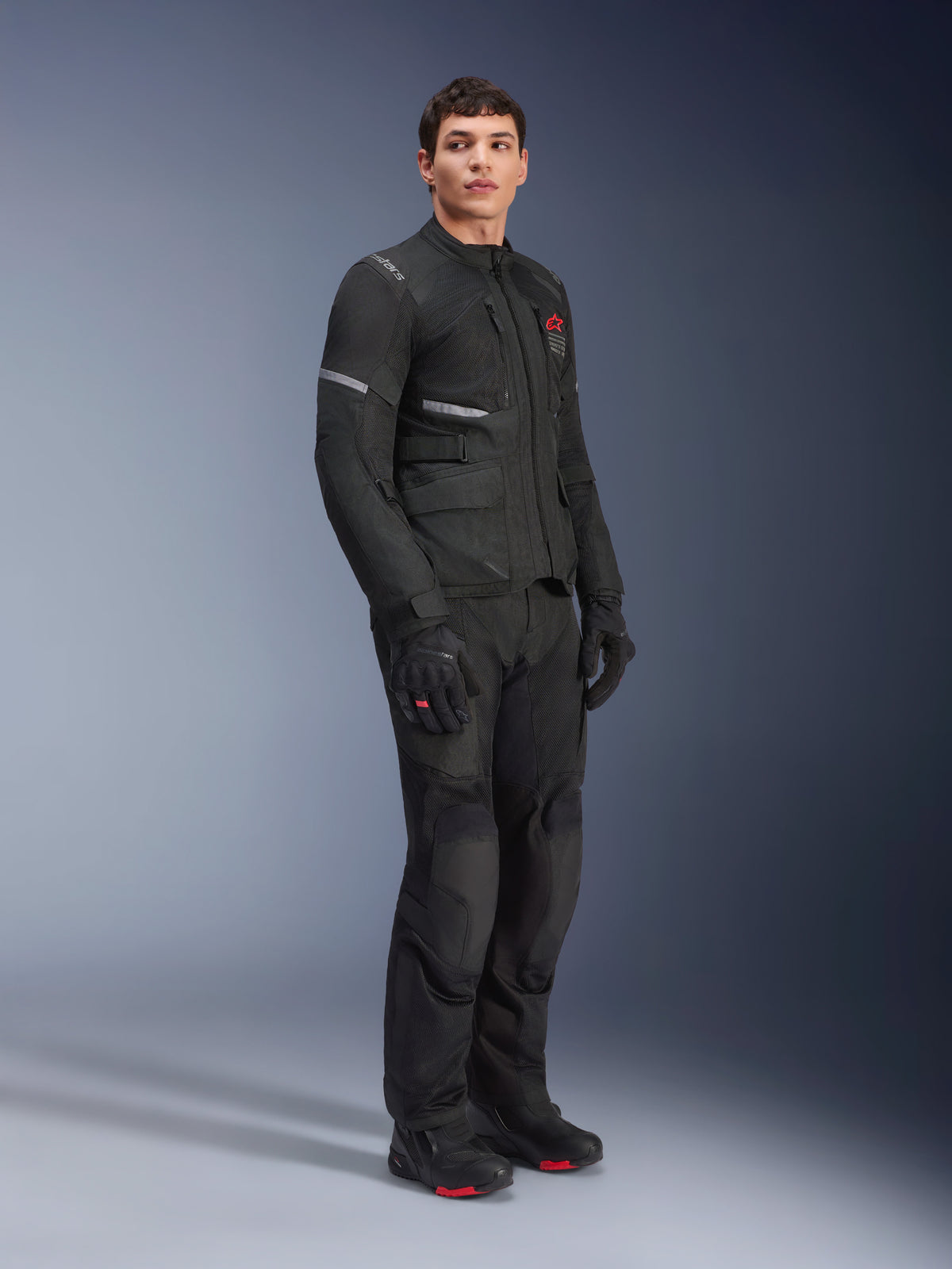 Giacca Alpinestars Andes Air Drystar®, nera, indossata da un modello come parte di un outfit completo da moto adventure con pantaloni, guanti e stivali abbinati, con pannelli di ventilazione in rete, logo rosso sul petto e strisce riflettenti per la visibilità touring.