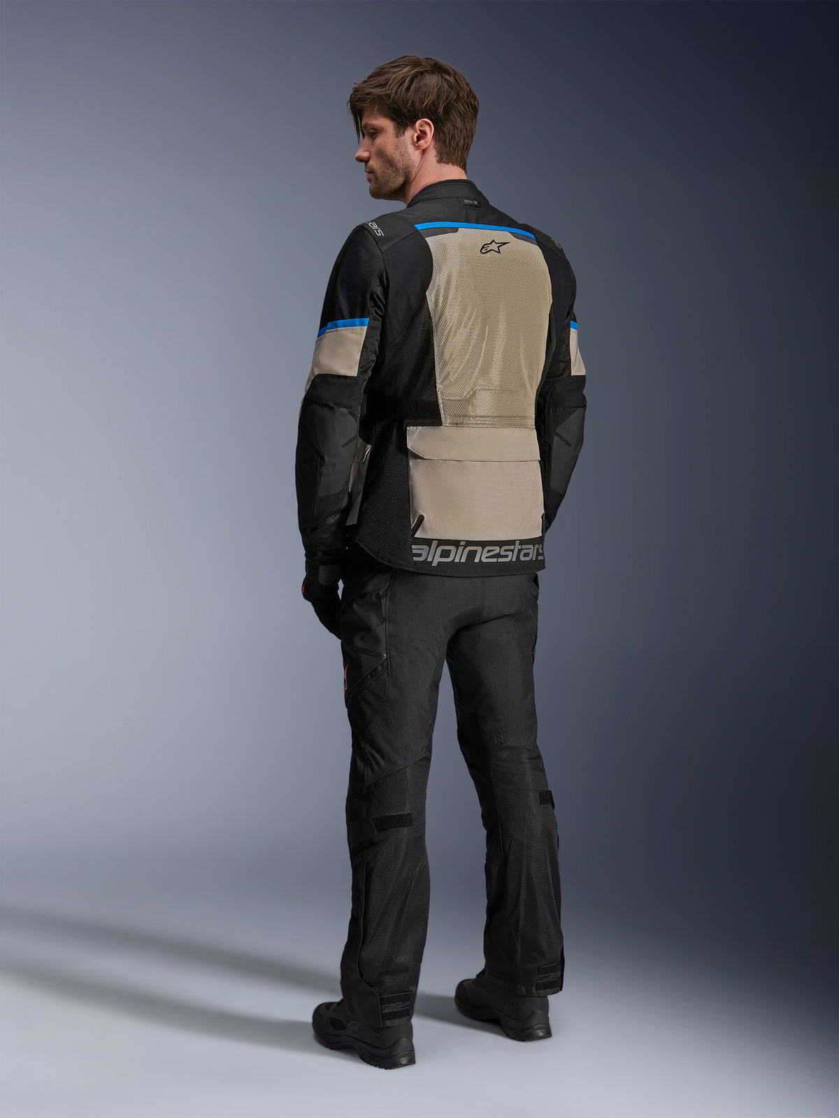 Giacca Alpinestars Andes Air Drystar®, nero, grigio Laurel e blu acceso, vista posteriore che mostra l'ampio pannello di ventilazione in rete sulla schiena, logo sul bordo inferiore, indossata con pantaloni da moto e stivali neri.