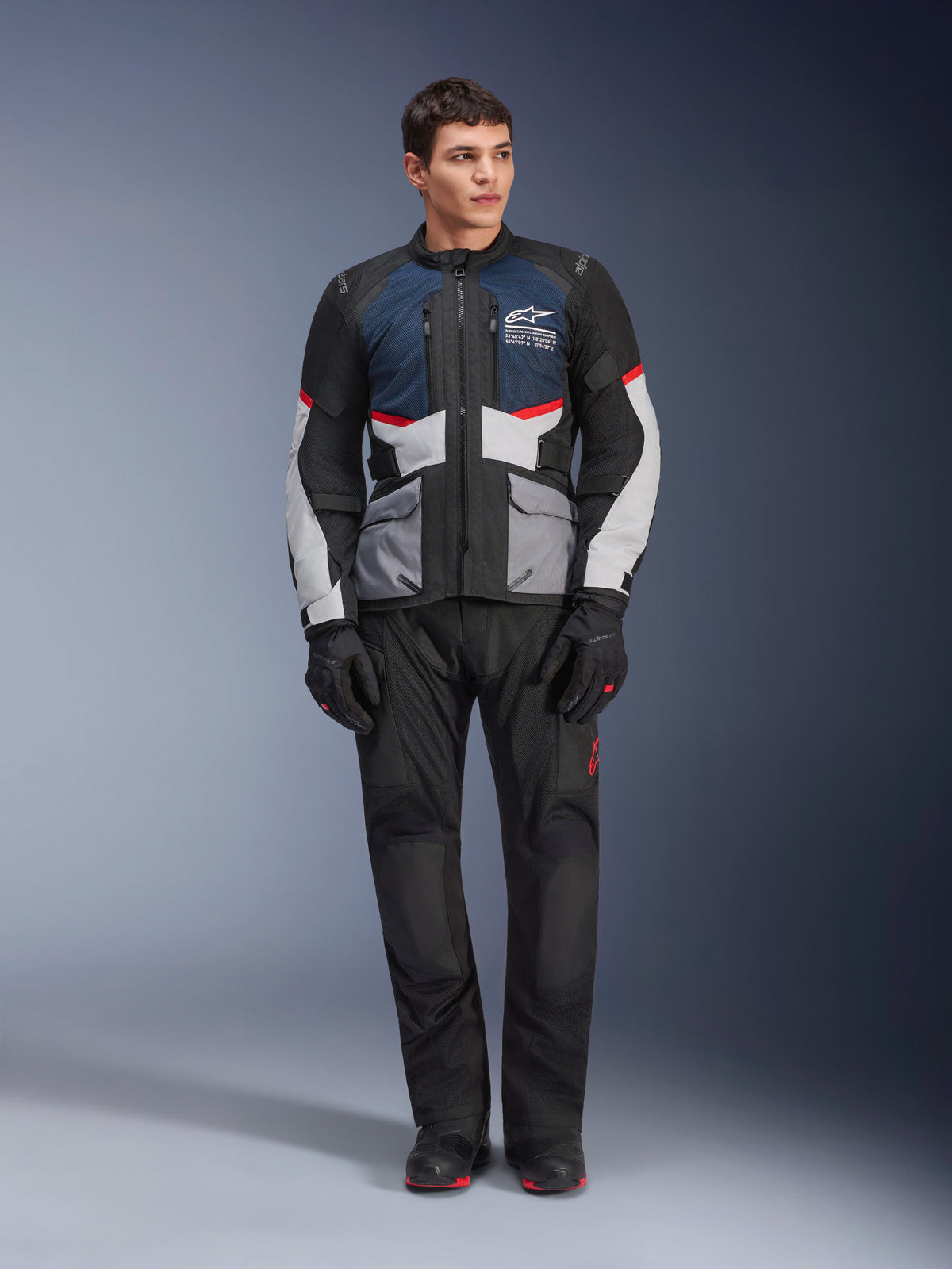 Giacca Alpinestars Andes Air Drystar®, giacca da moto touring, blu scuro, nero e grigio ghiaccio, indossata con pantaloni e guanti adventure neri coordinati, con pannelli in mesh per il flusso d'aria e un design tecnico multitasche.