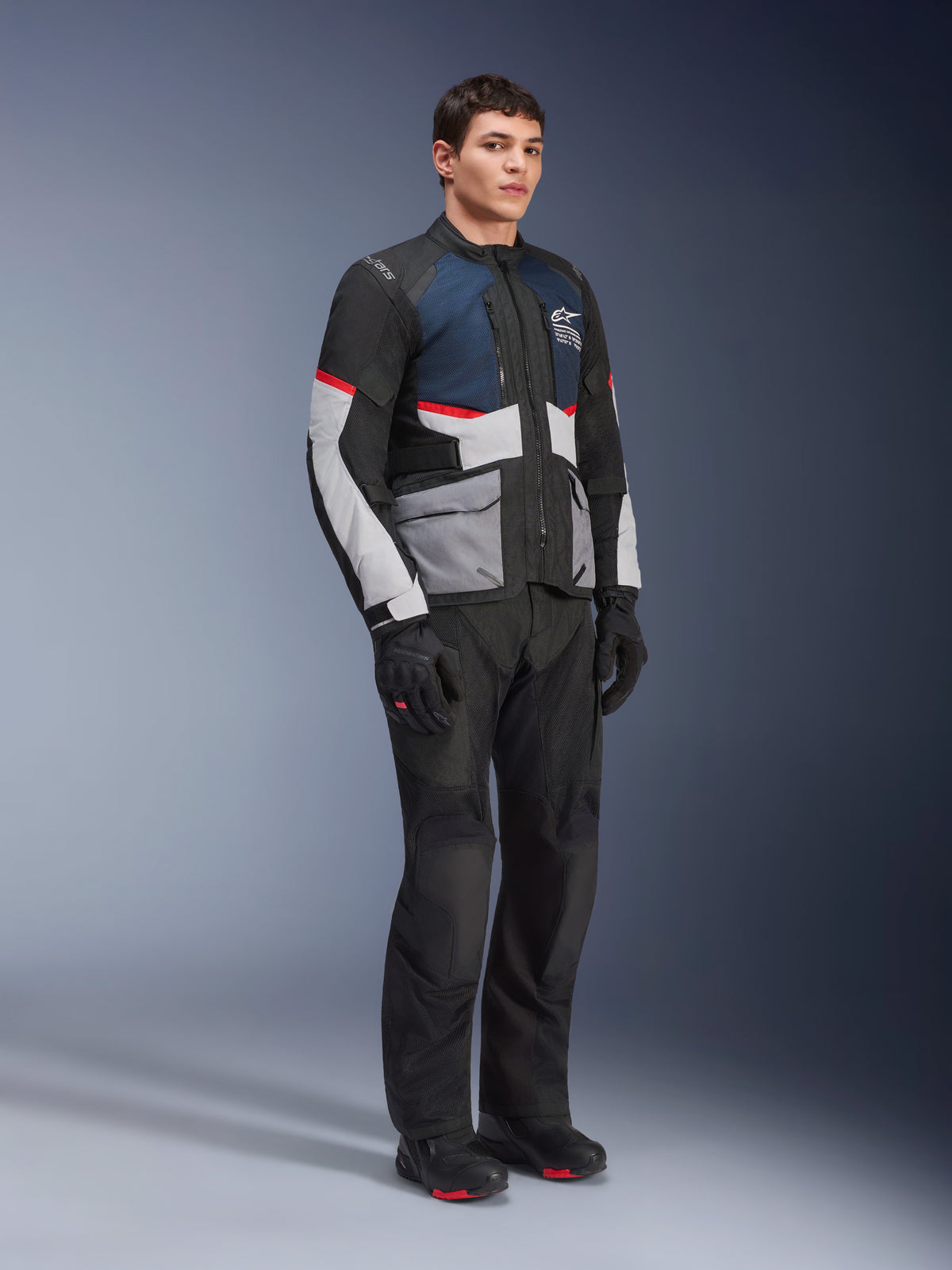 Modello che indossa la giacca Alpinestars Andes Air Drystar® in Deep Blue Black Ice Gray, abbinata a pantaloni, guanti e stivali da moto neri, con ampi pannelli in rete per la ventilazione, tasche grigio chiaro e accenti rossi, progettata per l'adventure touring.